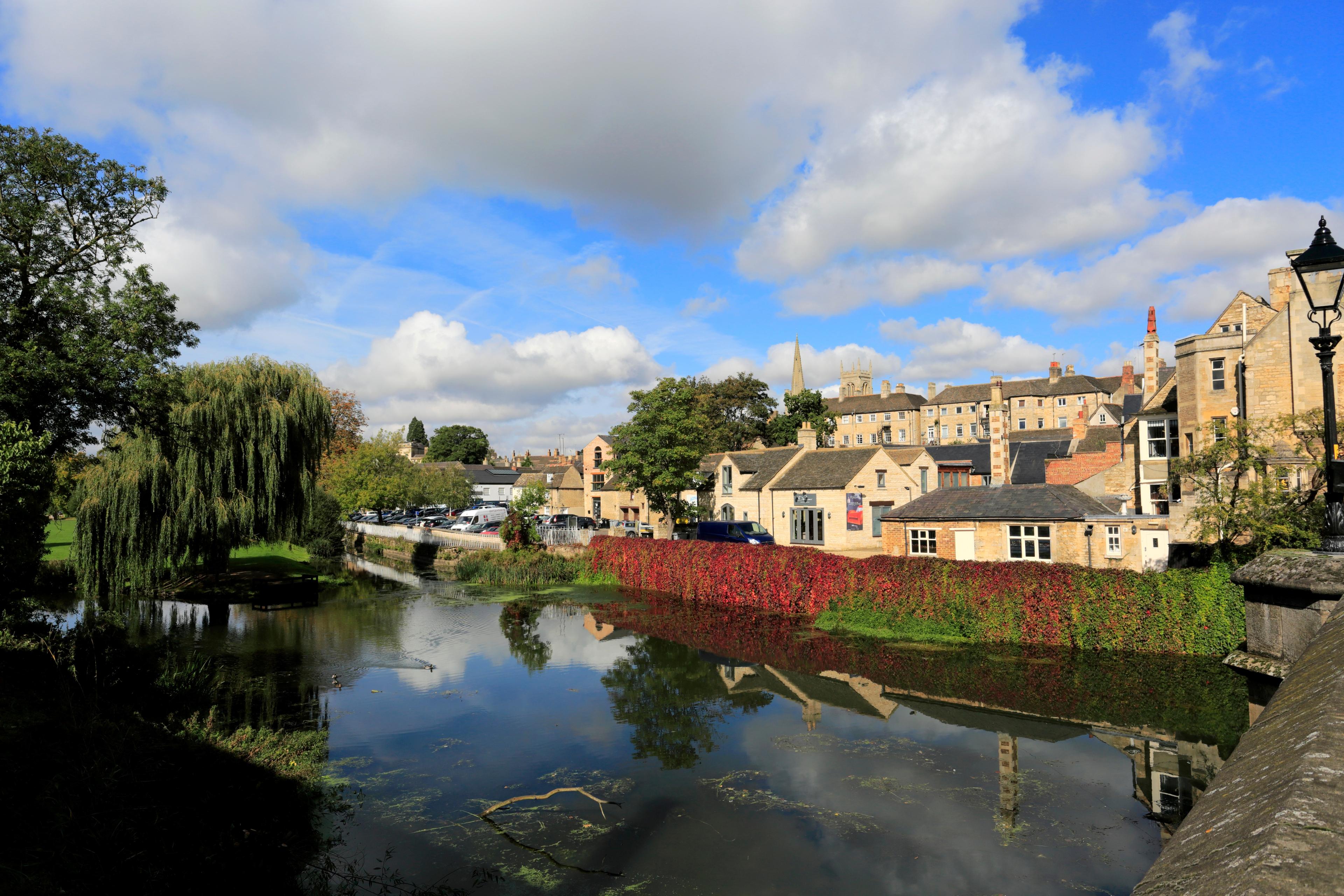 Quaint village scene with stone buildings, a tree-lined river, and a bright sky with fluffy clouds reflecting in the water.