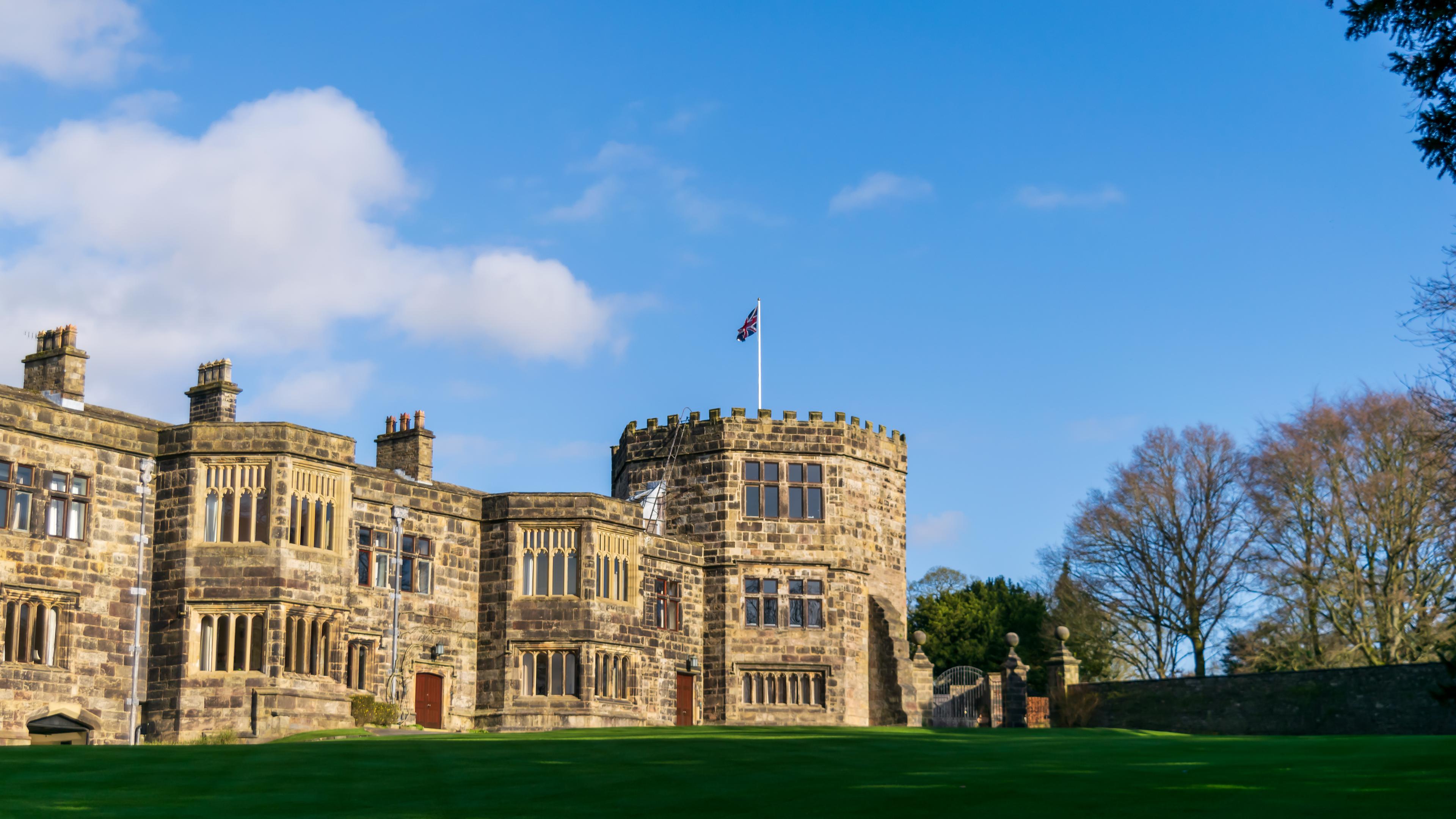 A historic stone castle with a round tower and the British flag, set against a blue sky with clouds and surrounded by trees and a manicured lawn.