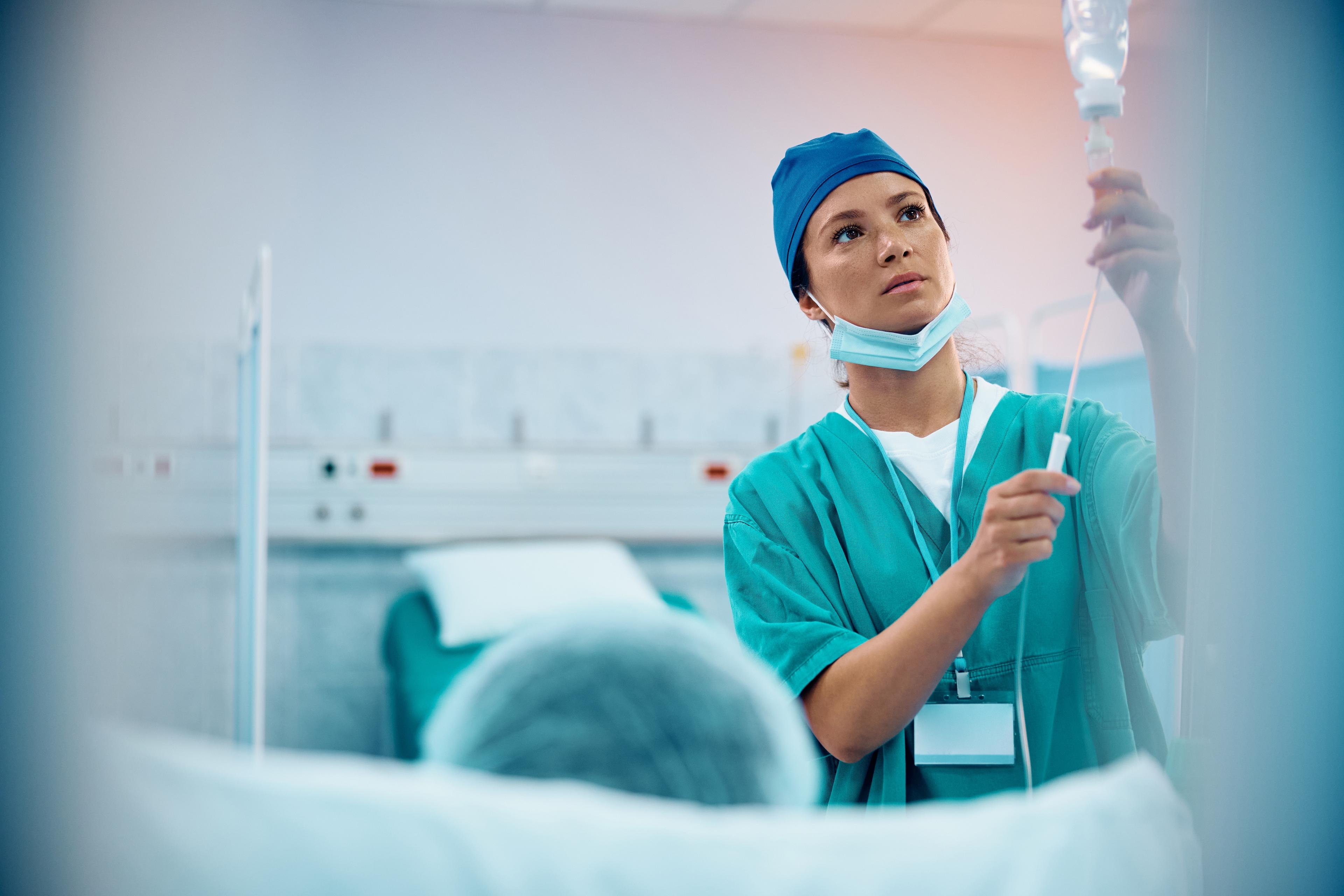 A nurse in scrubs and a cap adjusts an IV drip in a hospital room, with a patient lying in the foreground.