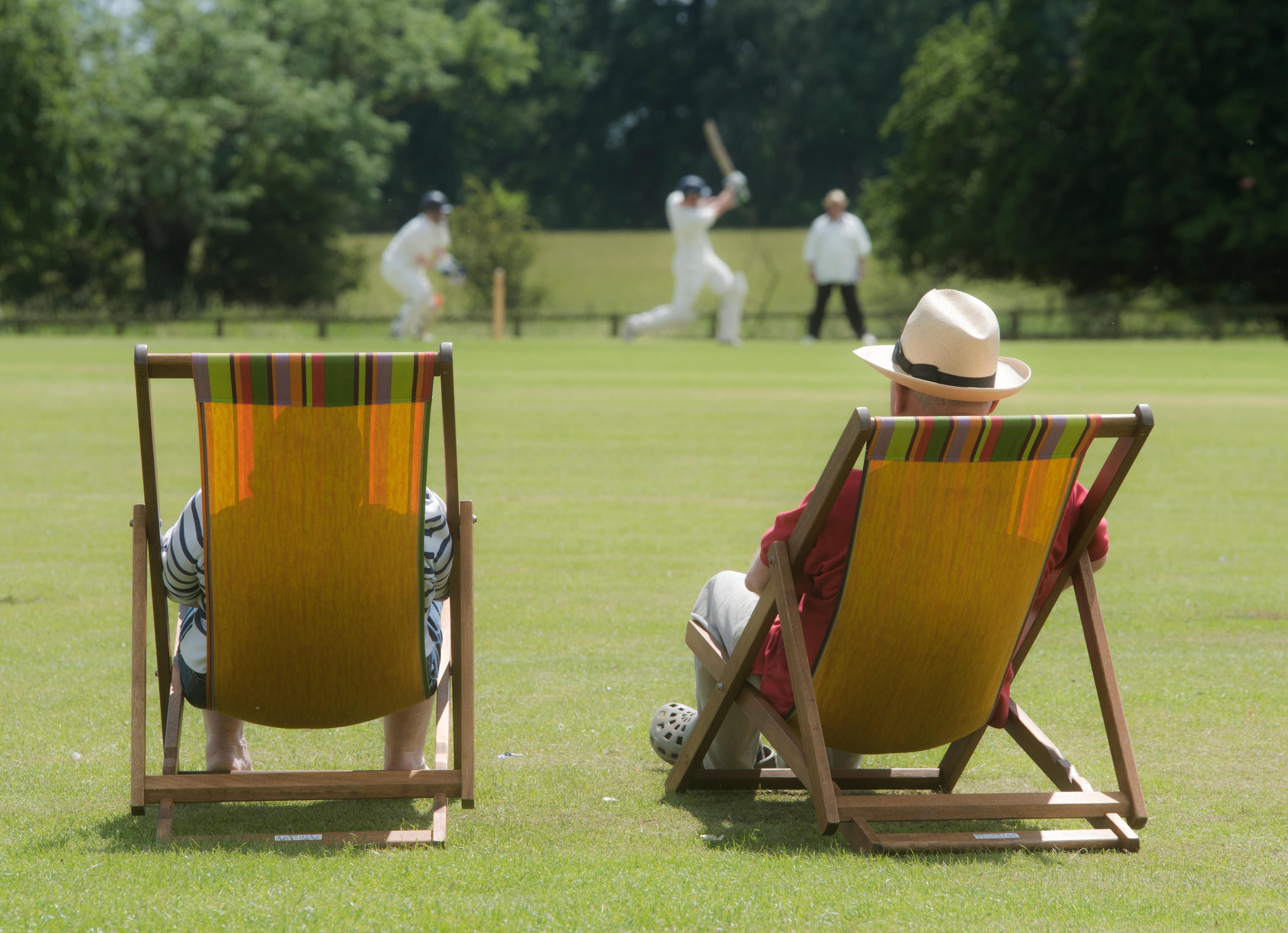 Two people sit in deck chairs watching a cricket match on a sunny day, with players in white uniforms in the background.