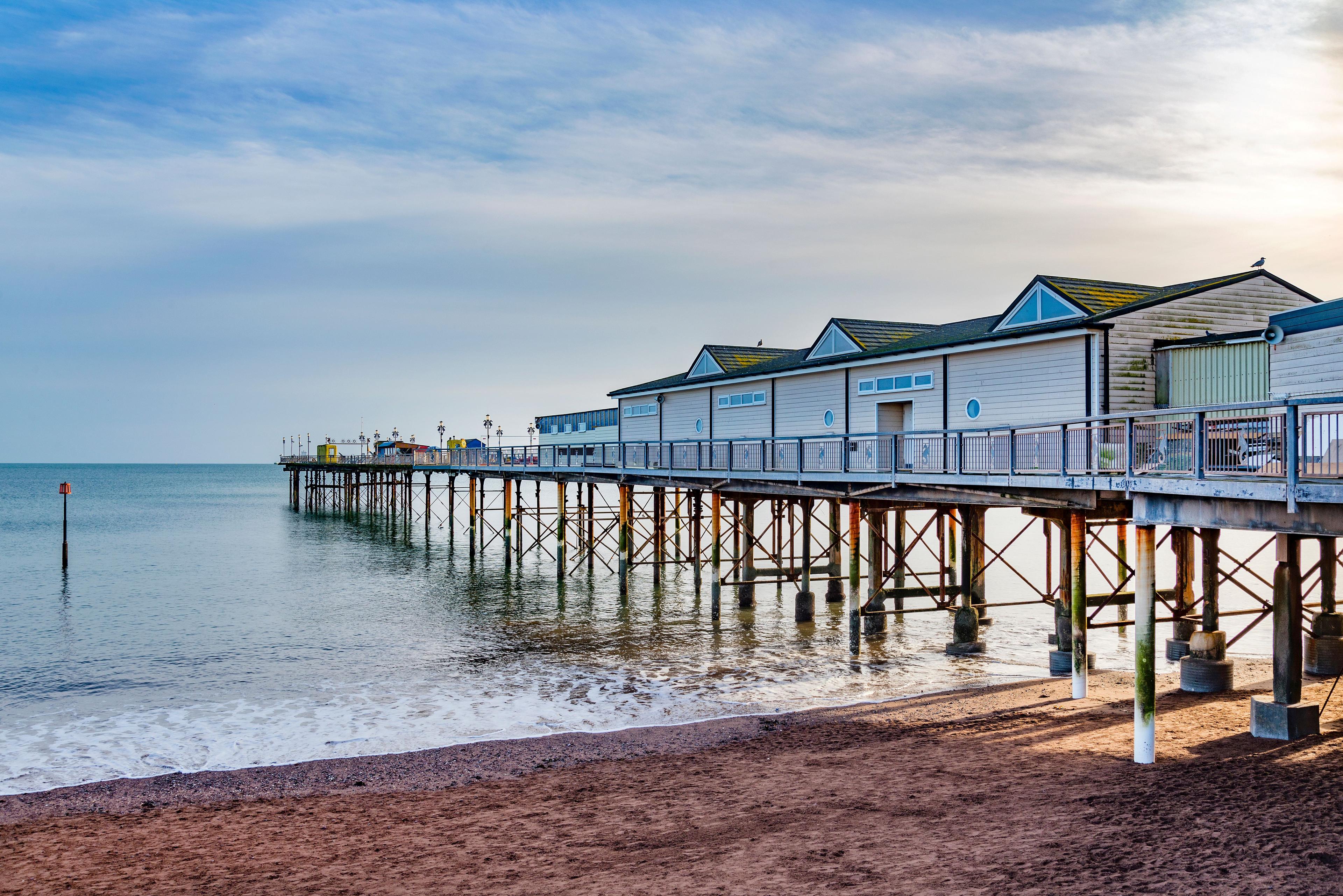 White-and-blue beach huts line a long seaside pier over calm water, beneath a cloudy sky and beside a pebbly shore, creating a tranquil coastal scene.