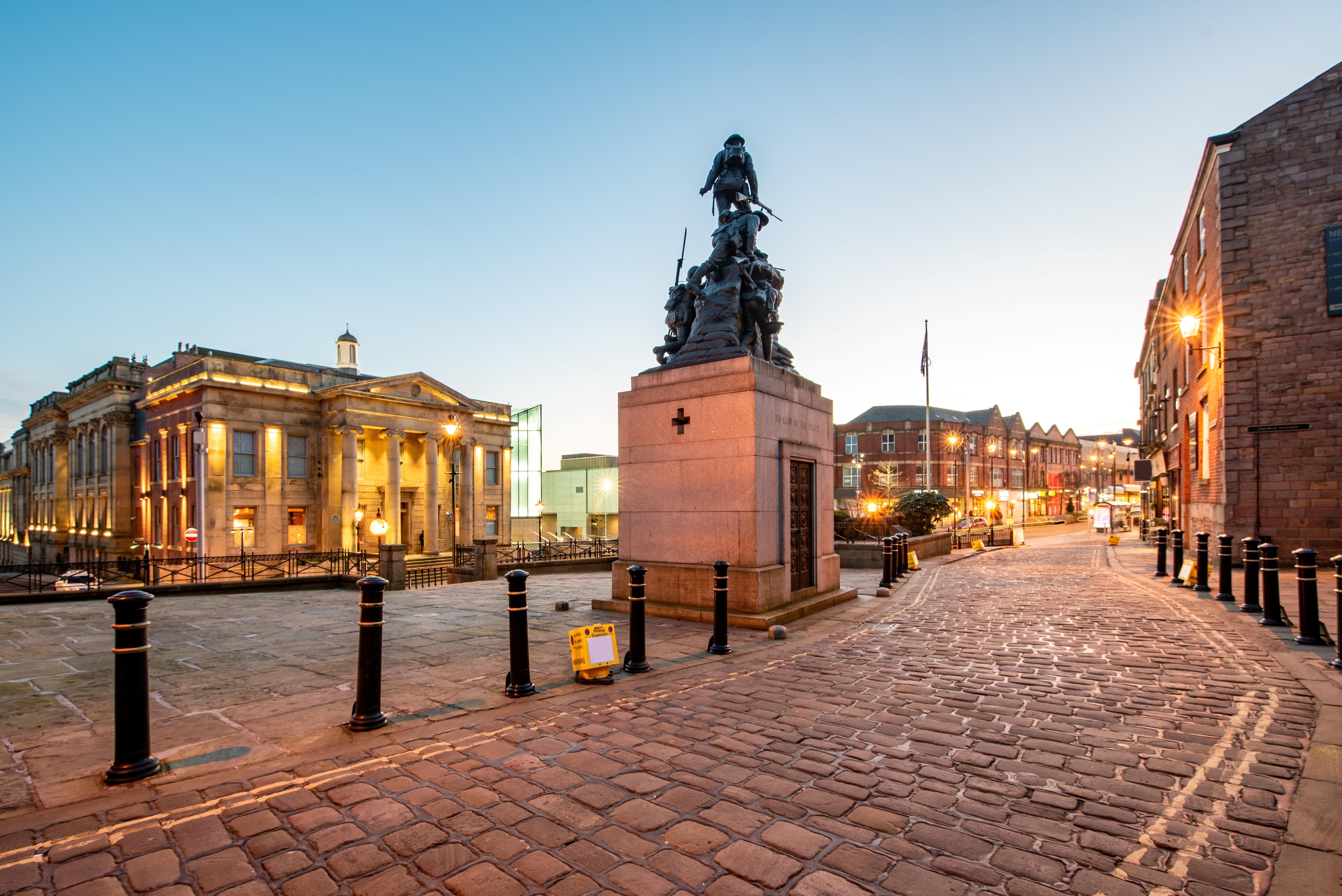 An illuminated war memorial sculpture stands in a cobbled square outside a grand historic town hall in Oldham, West Yorkshire, with surrounding stone buildings and streetlights glowing at dusk.