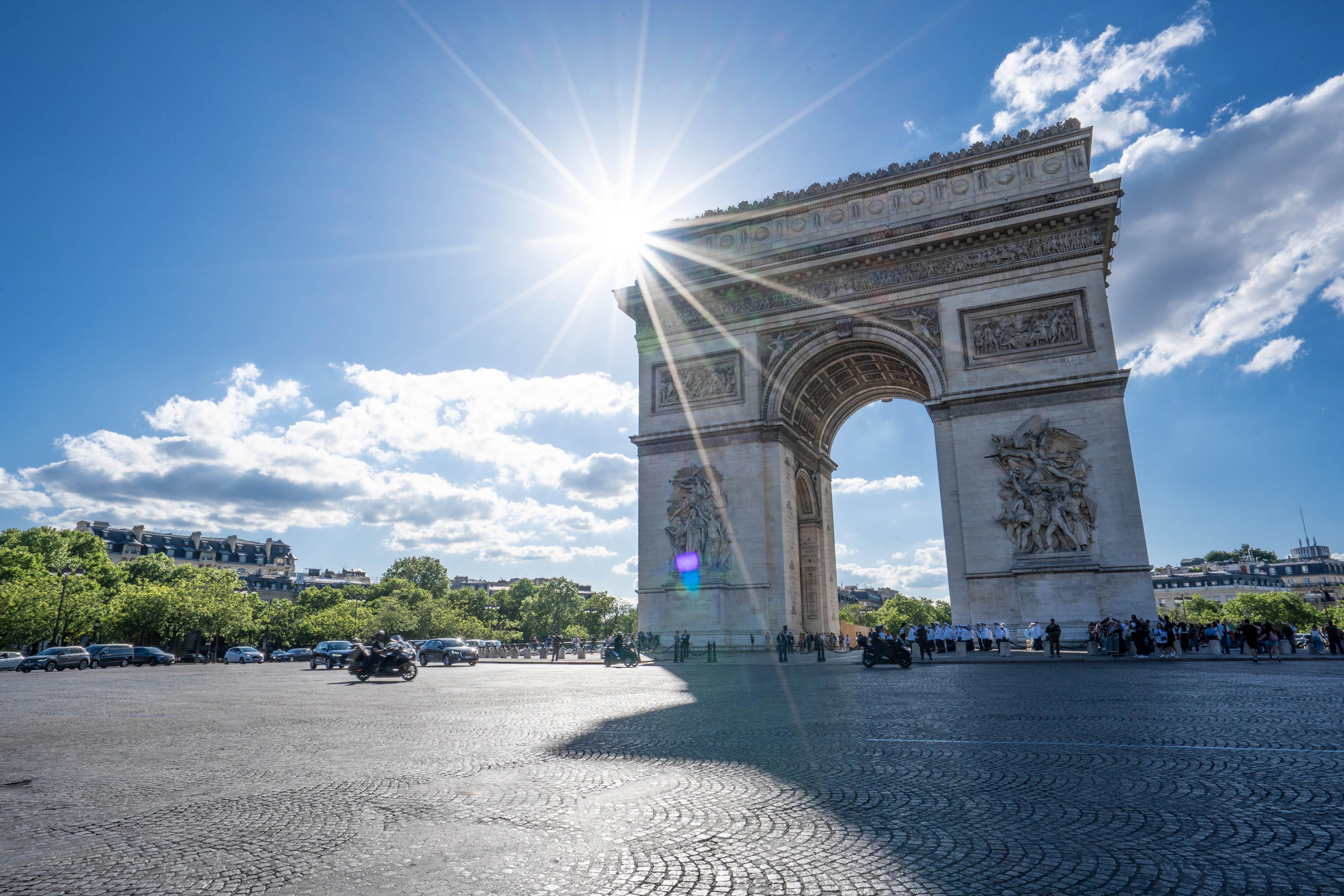 The Arc de Triomphe in Paris under a bright blue sky, with sunbeams shining above and some clouds in the distance.