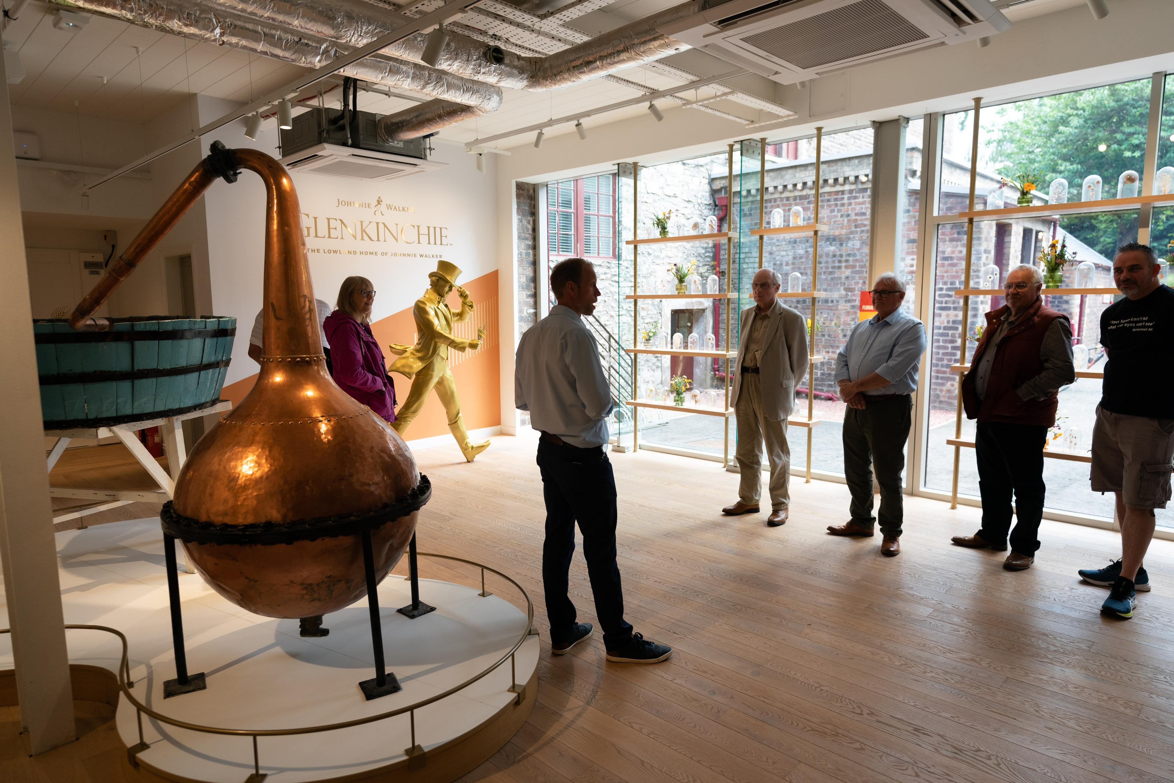 A tour group in a distillery room with a copper still, displayed under warm lighting, and a Johnnie Walker statue in the background.