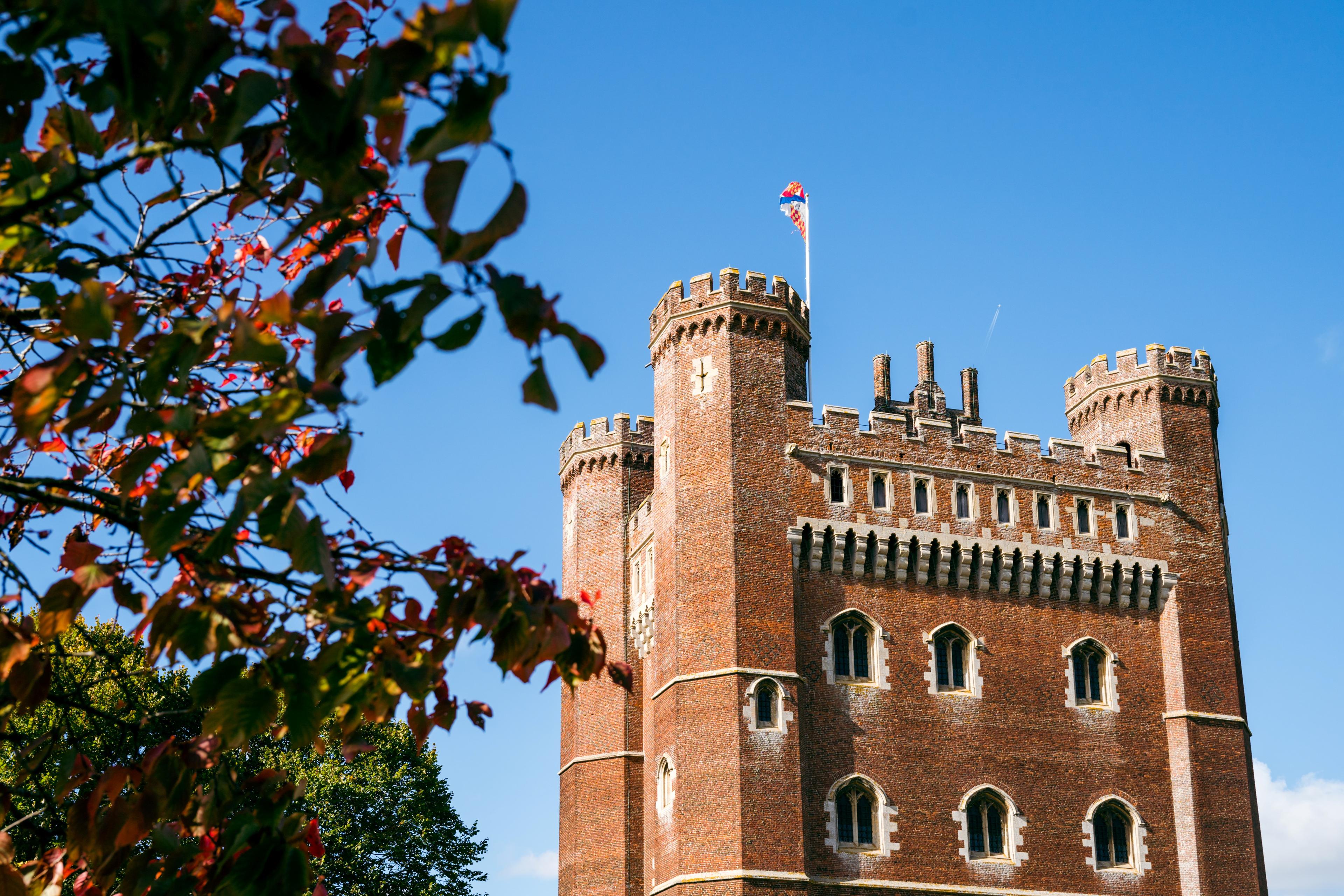 Brick castle tower with crenellations and flag, set against a clear blue sky. Autumn leaves frame the left side of the image.