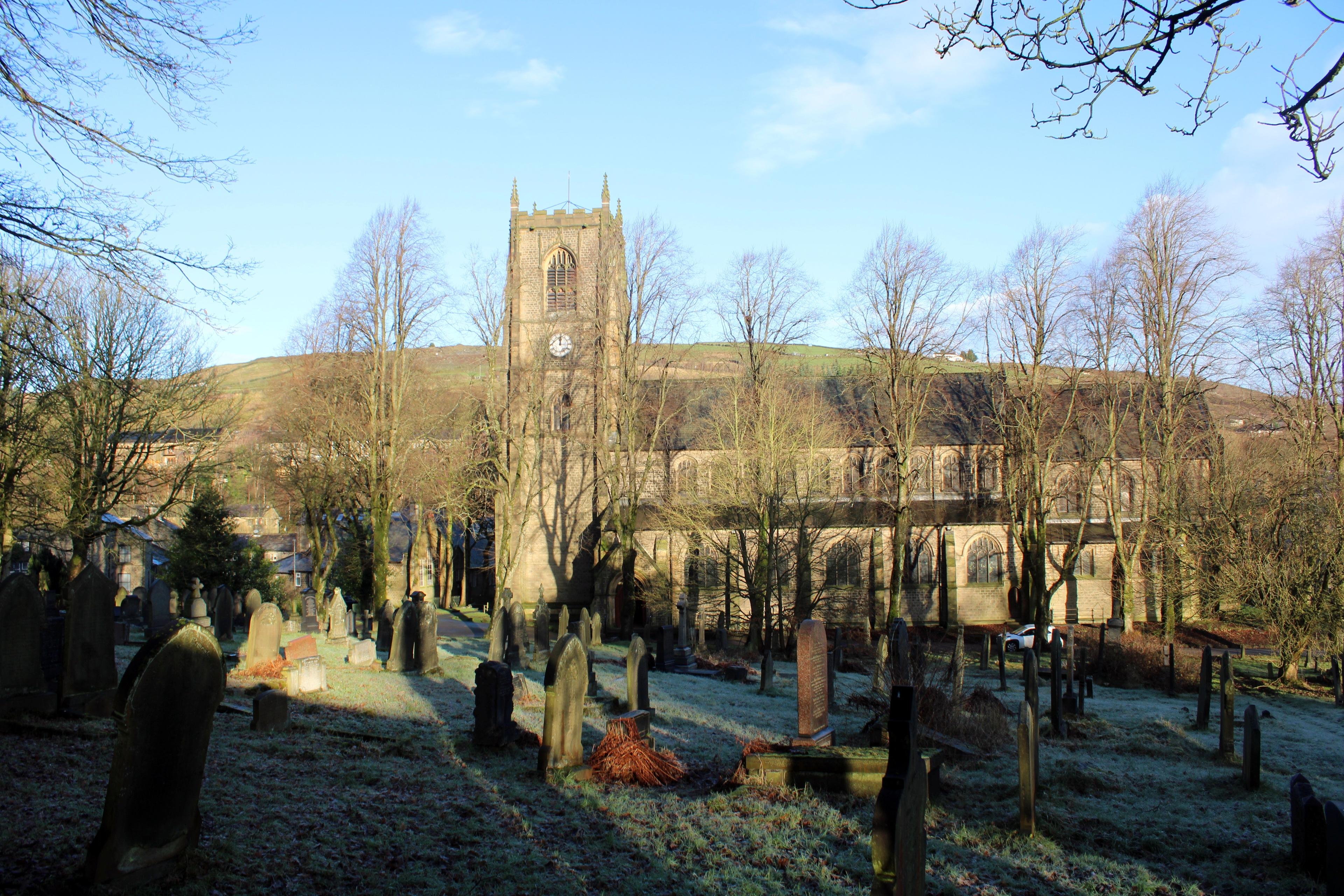 St Bartholomew’s Church in Marsden, West Yorkshire, viewed from its frosty churchyard, with a tall stone tower, bare trees, gravestones, and rolling hills in the background beneath a clear blue sky.