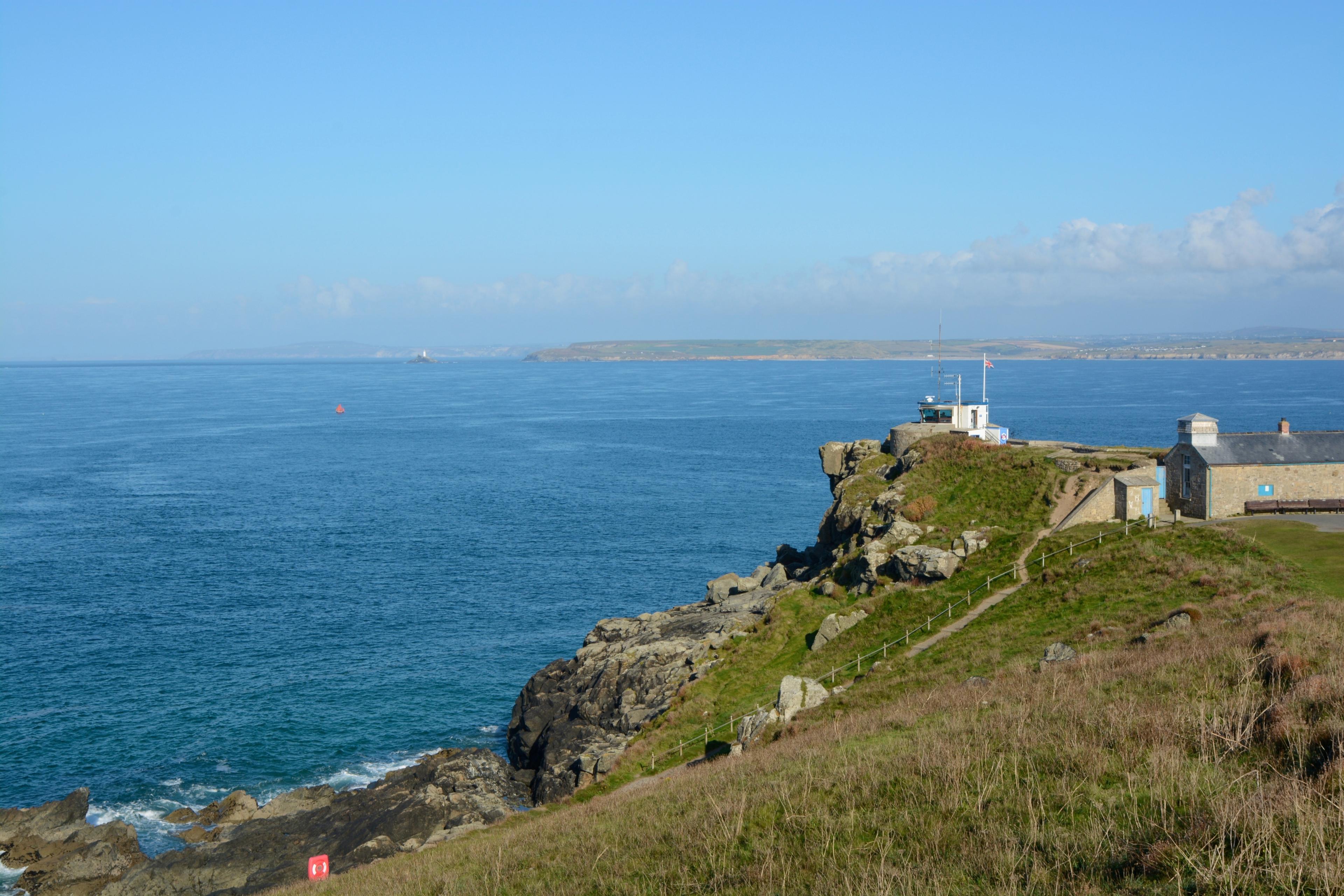 Clifftop station overlooking a vast blue sea with distant coastline under a clear sky. Rocky foreground with grassy path leading to buildings.