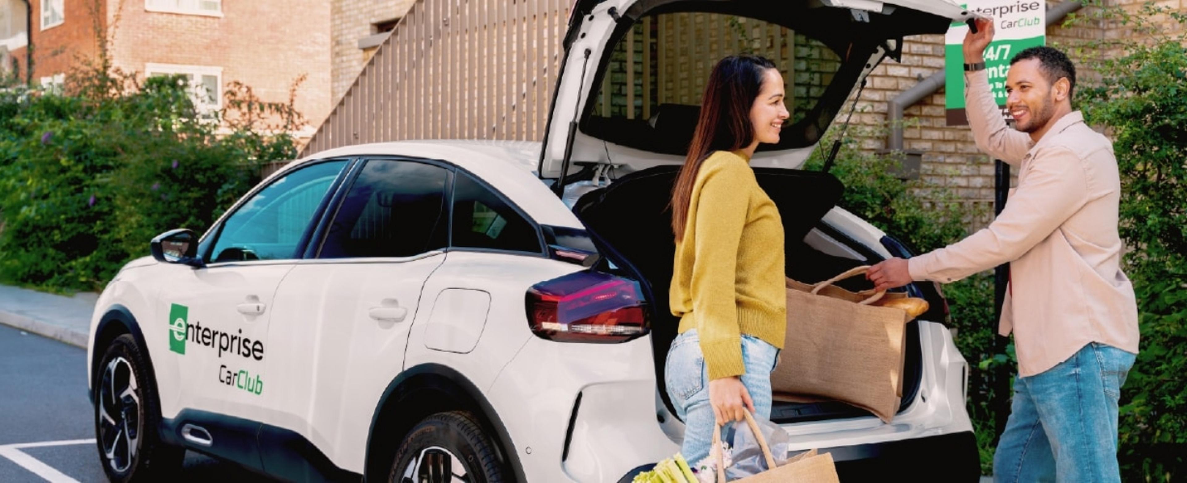 A man and woman load items into the trunk of a white Enterprise Car Club vehicle parked in a residential area labeled "Car Club."