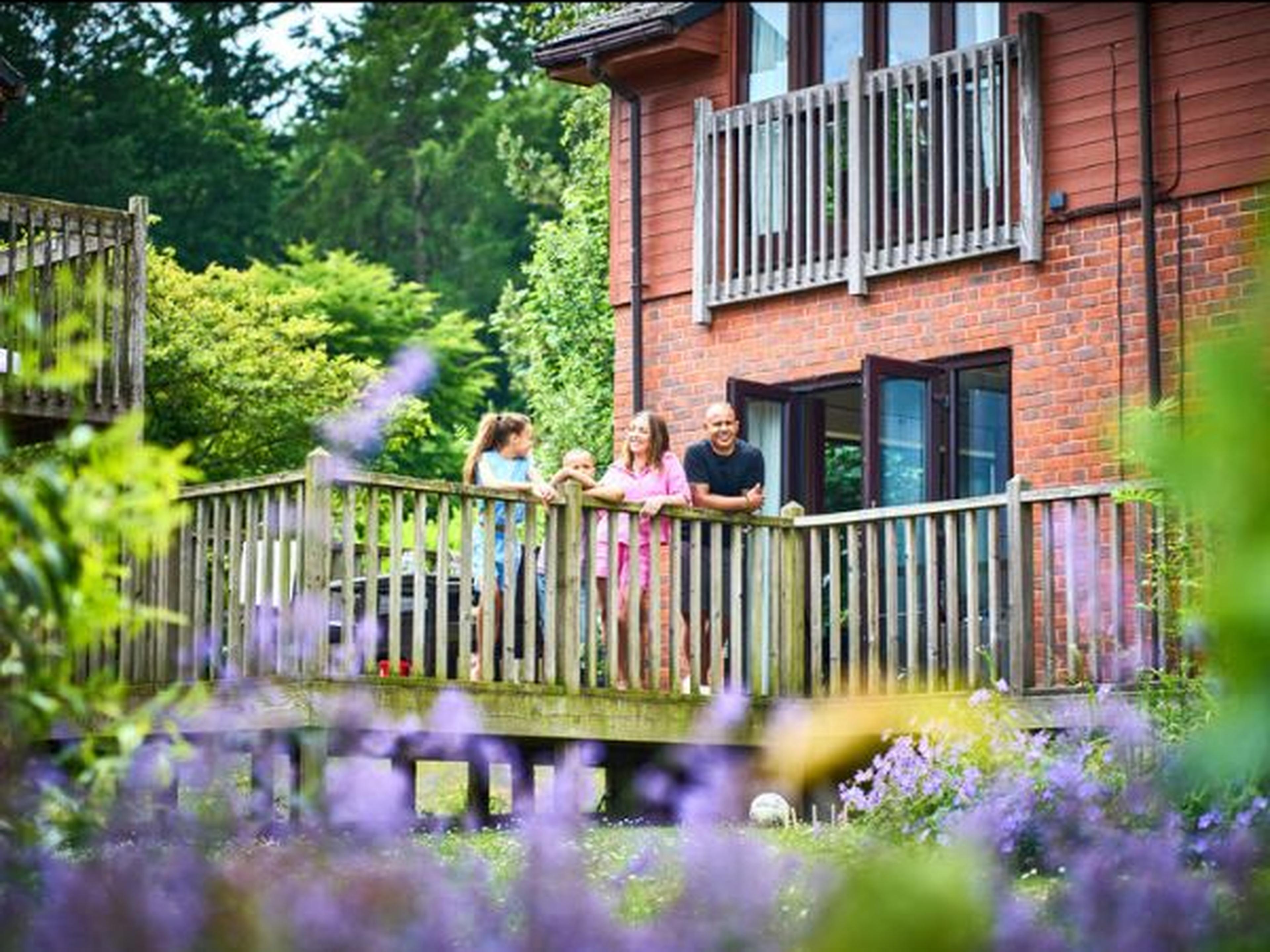 A family of four stands on a wooden deck outside a brick house, with lush greenery and purple flowers in the foreground.