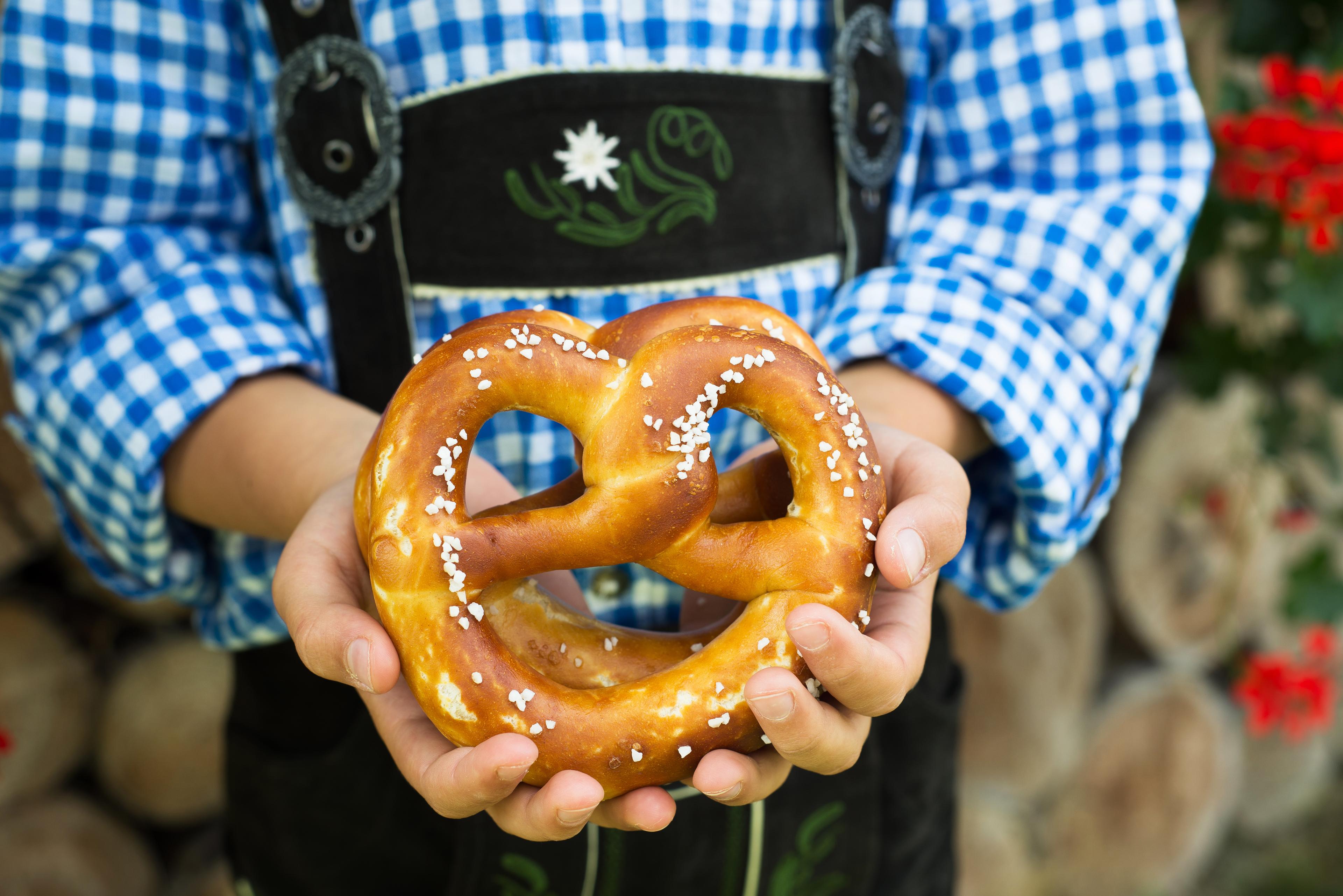 Person in blue checkered shirt holding a salted pretzel in front of a wooden background with red flowers.