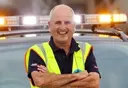 Smiling man in a safety vest stands with arms crossed in front of a vehicle with flashing lights.