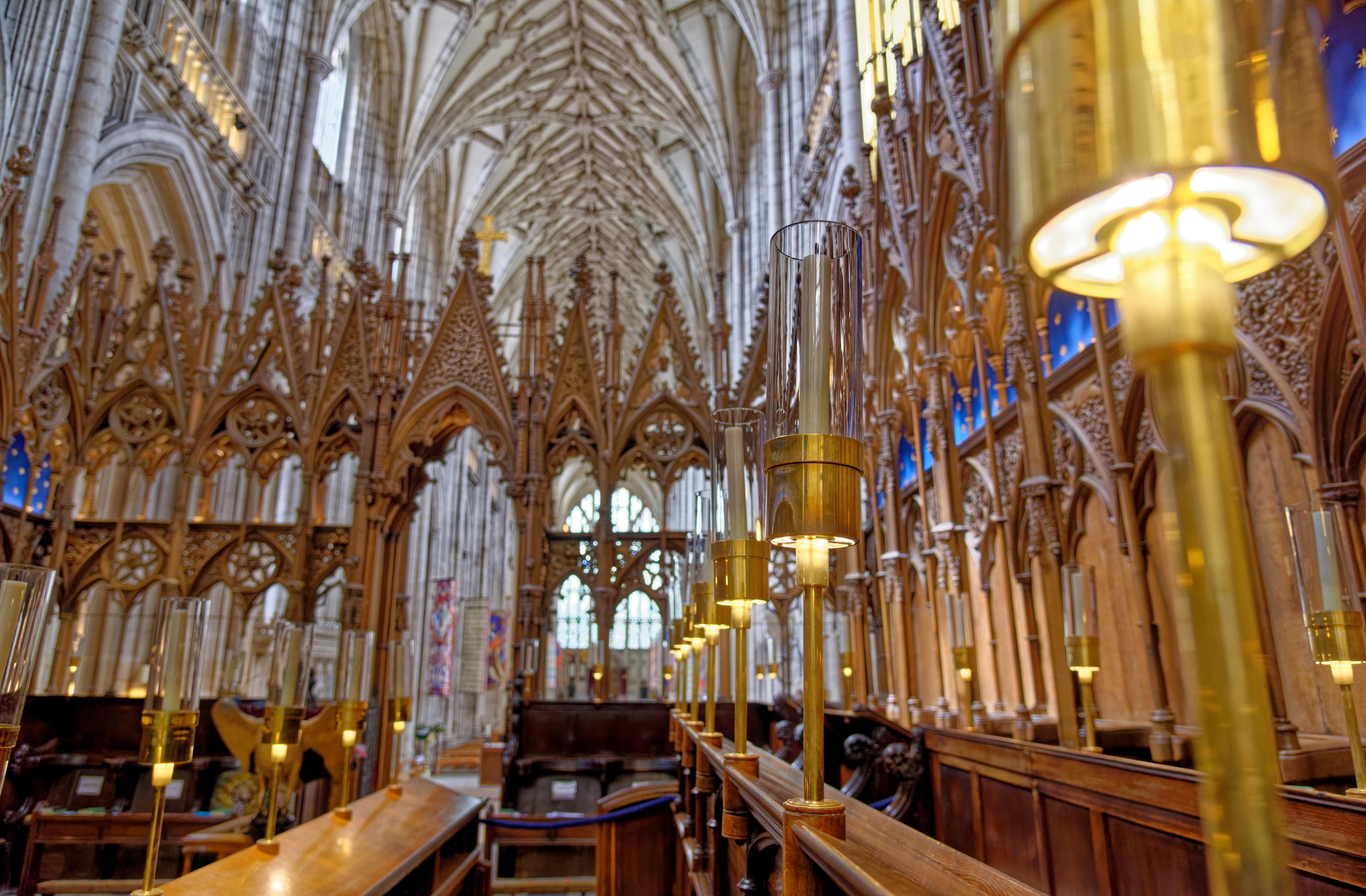 Intricate Gothic cathedral interior with vaulted ceilings, ornate wooden choir stalls, and tall candleholders emitting a warm glow.