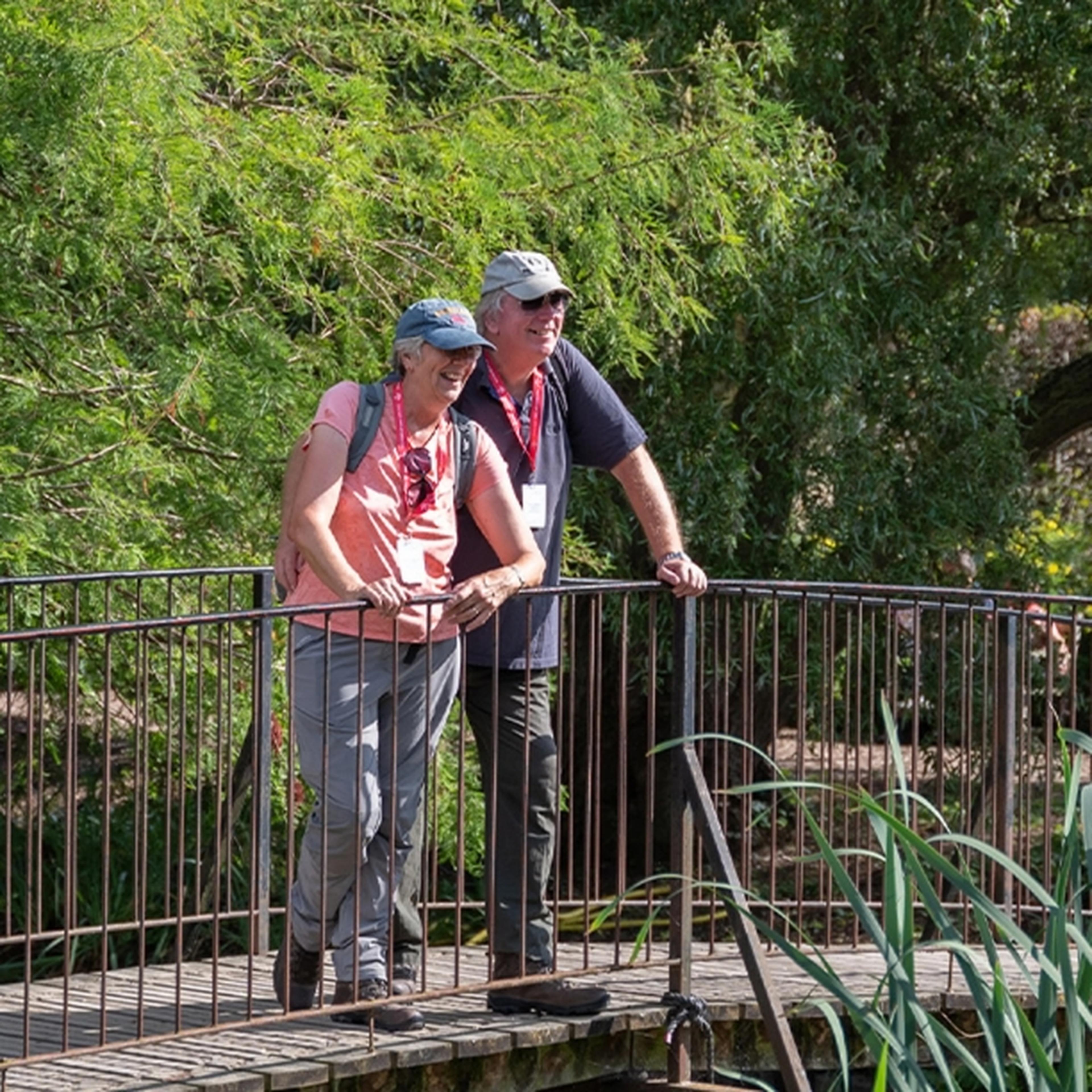 Two people wearing caps stand and lean on a curved railing, overlooking lush green foliage in a garden setting.