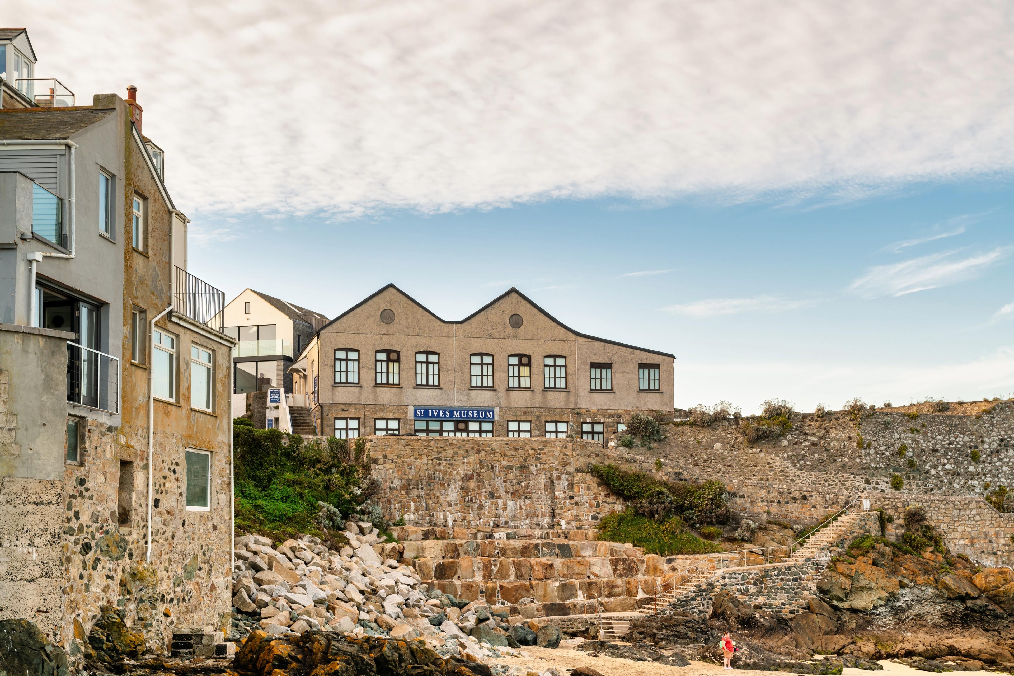 St Ives Museum on a rocky coastline, with adjacent buildings under a partly cloudy sky, and a person walking on the sandy beach below.