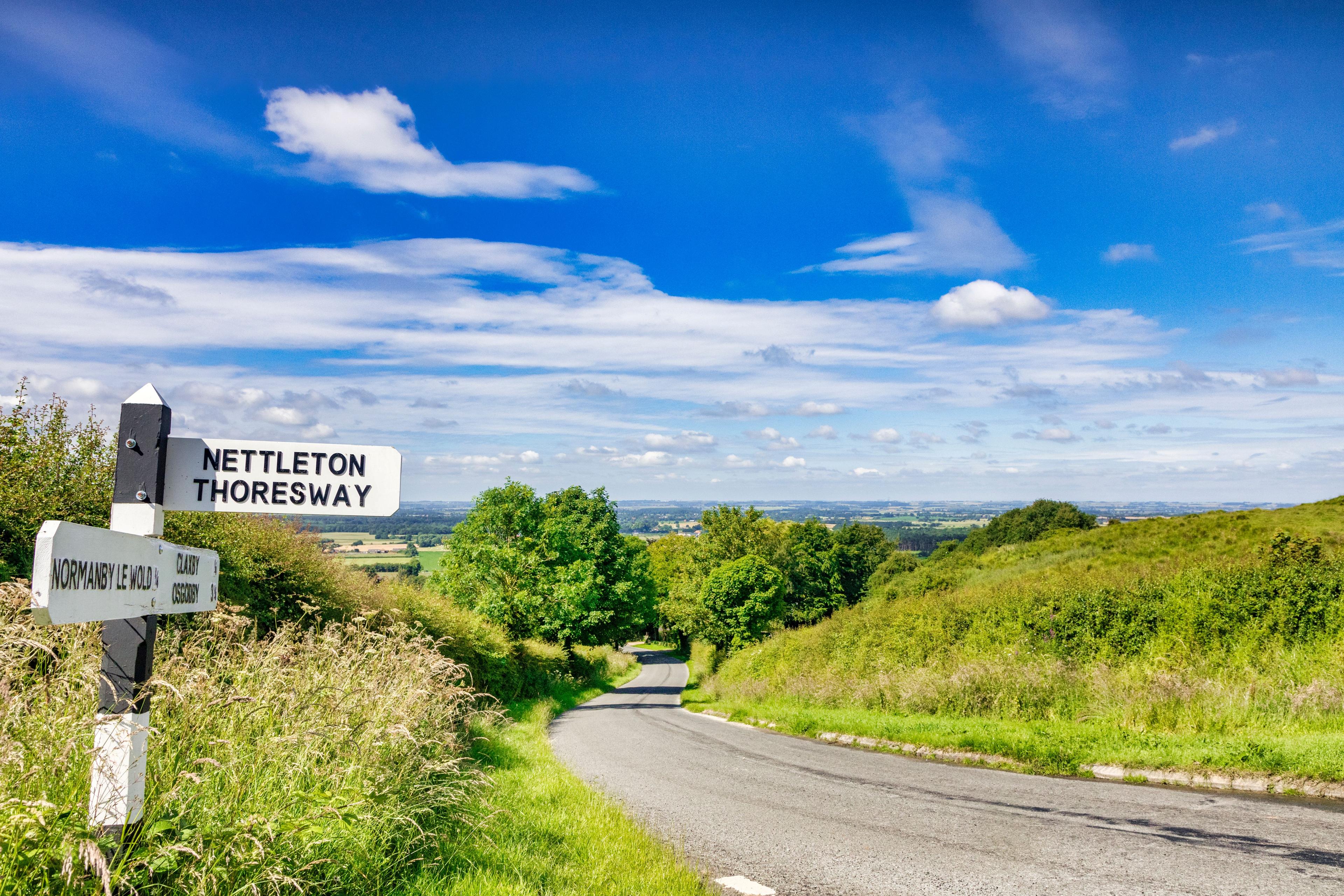 Country road with lush greenery and a signpost pointing to Nettleton and Thoresway under a bright blue sky with scattered clouds.