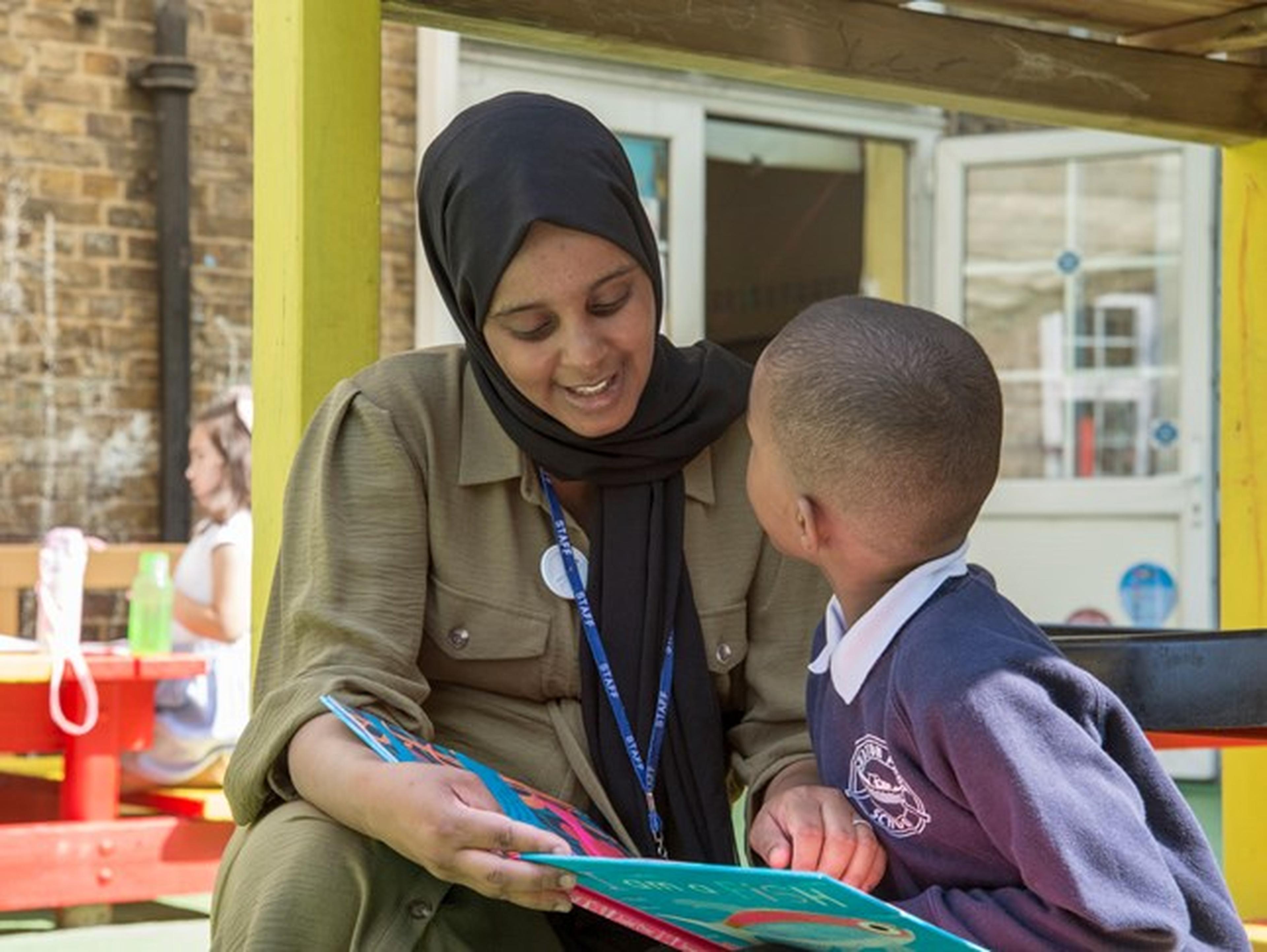 A woman  reads a colorful book to a young boy in a school uniform outside, both smiling and engaged.