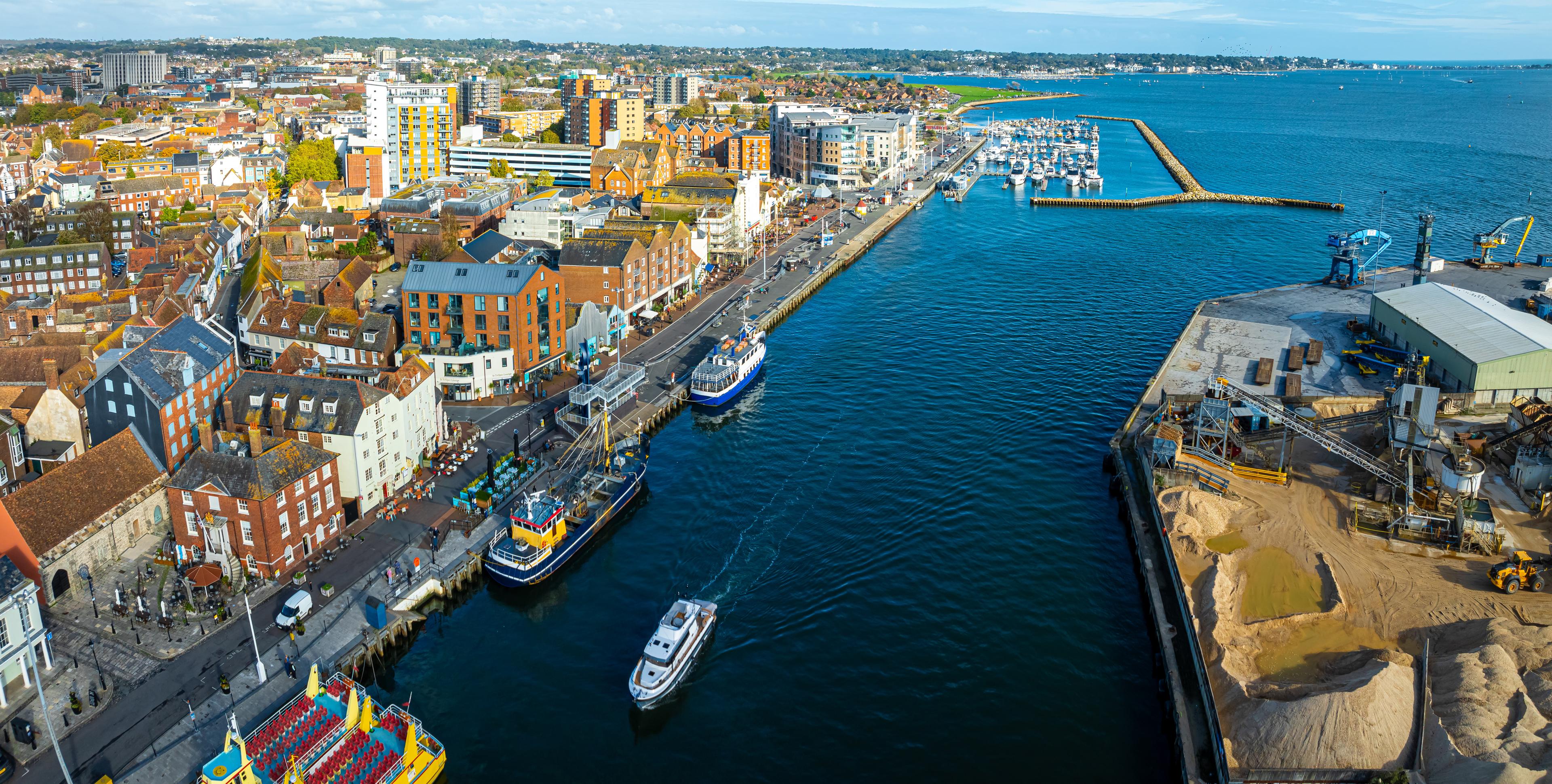 Aerial view of a coastal town with boats in a harbor, colorful buildings, and an industrial area with piles of sand.