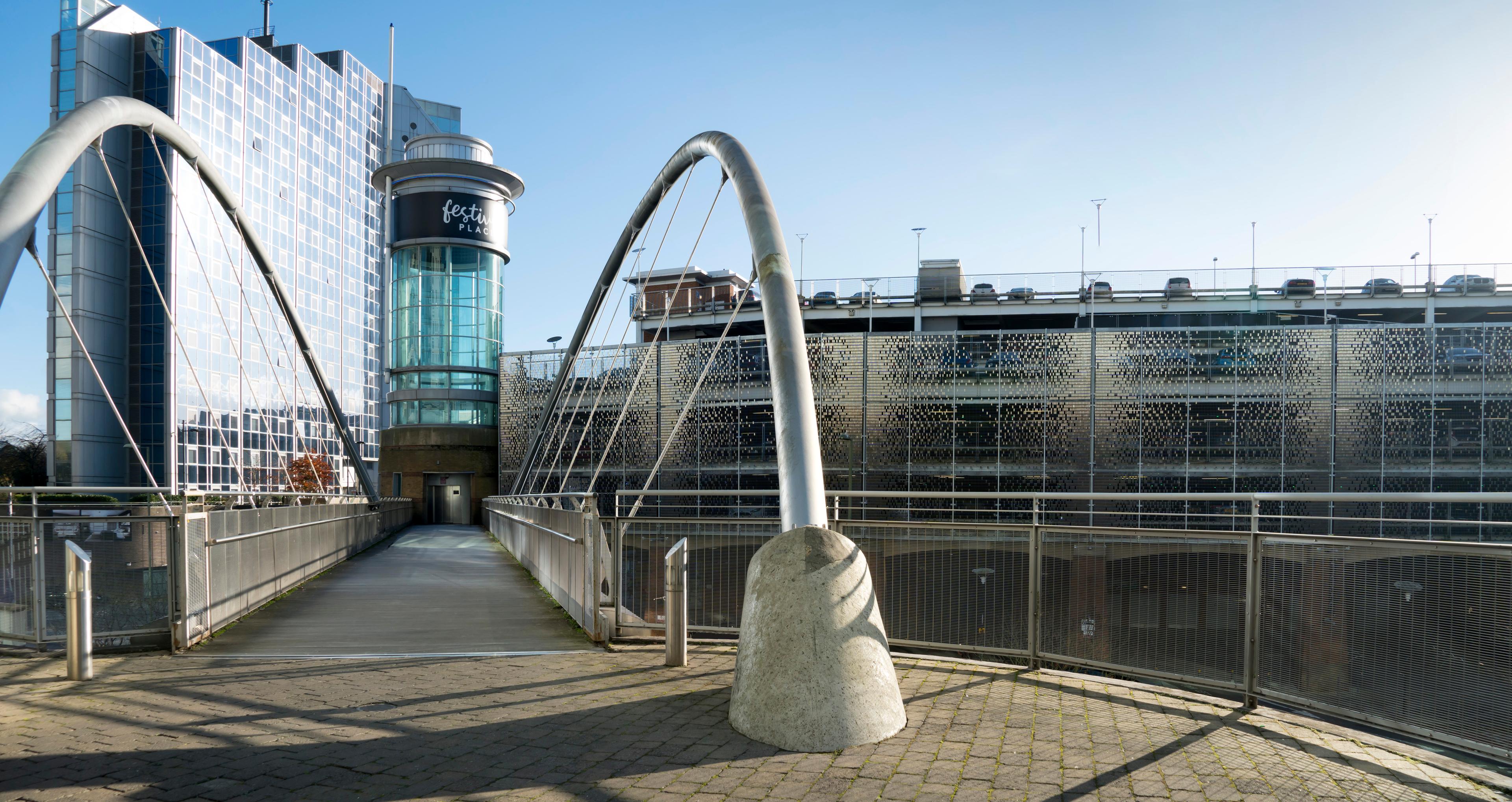 Modern pedestrian bridge with curved steel arches, leading to a glass building with a tall cylindrical section, under a clear blue sky.