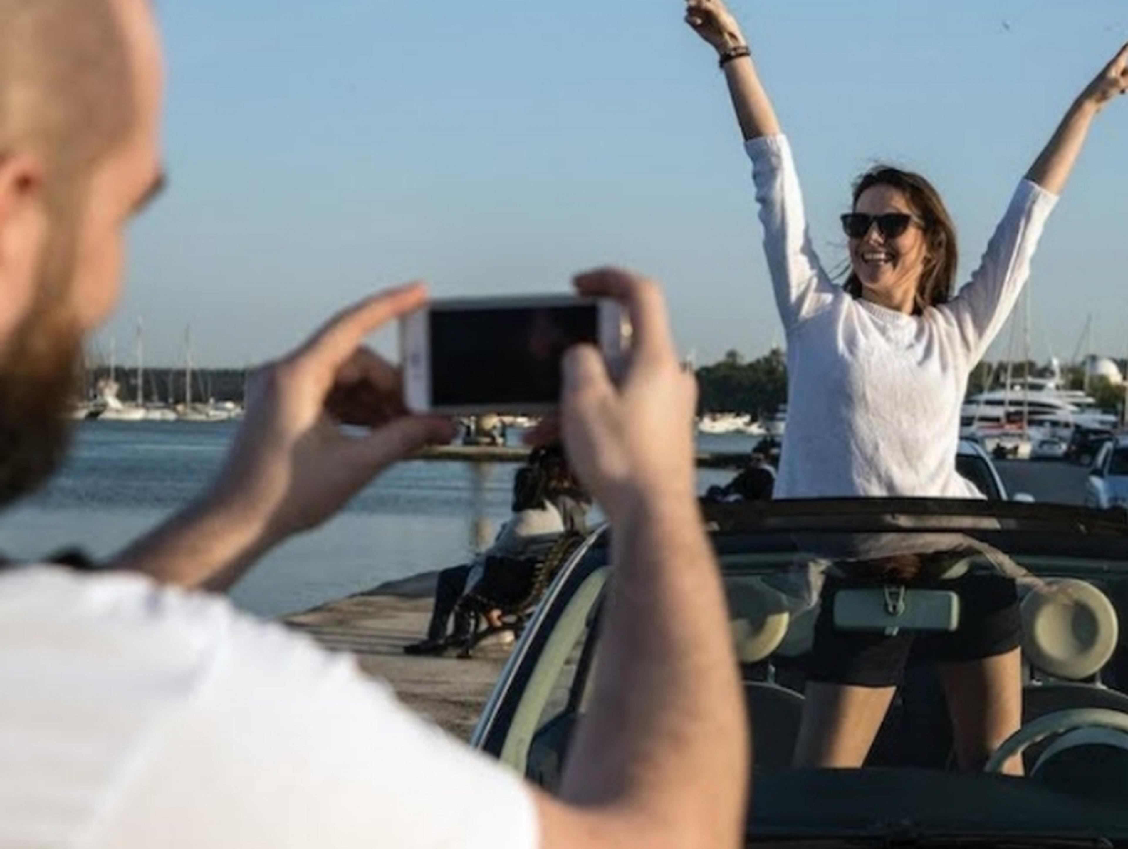 Man taking a photograph of a woman standing up in her car with arms aloft