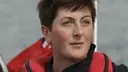 Person with short brown hair wearing a red life jacket, gazing ahead with a serious expression. Background shows a blurred flag and water.