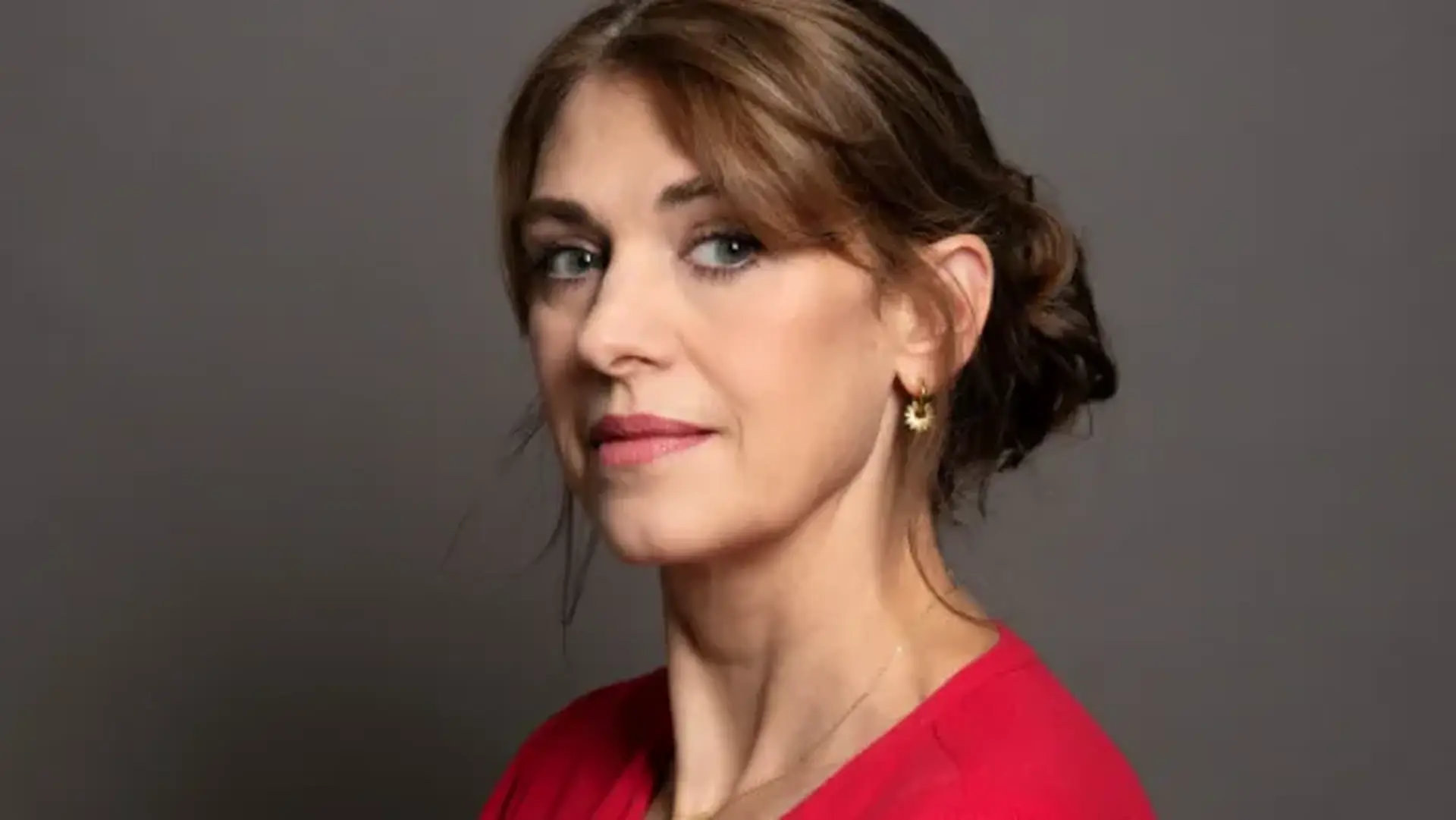 Woman with brown hair in an updo wearing a red top and gold earrings, posing against a gray background.