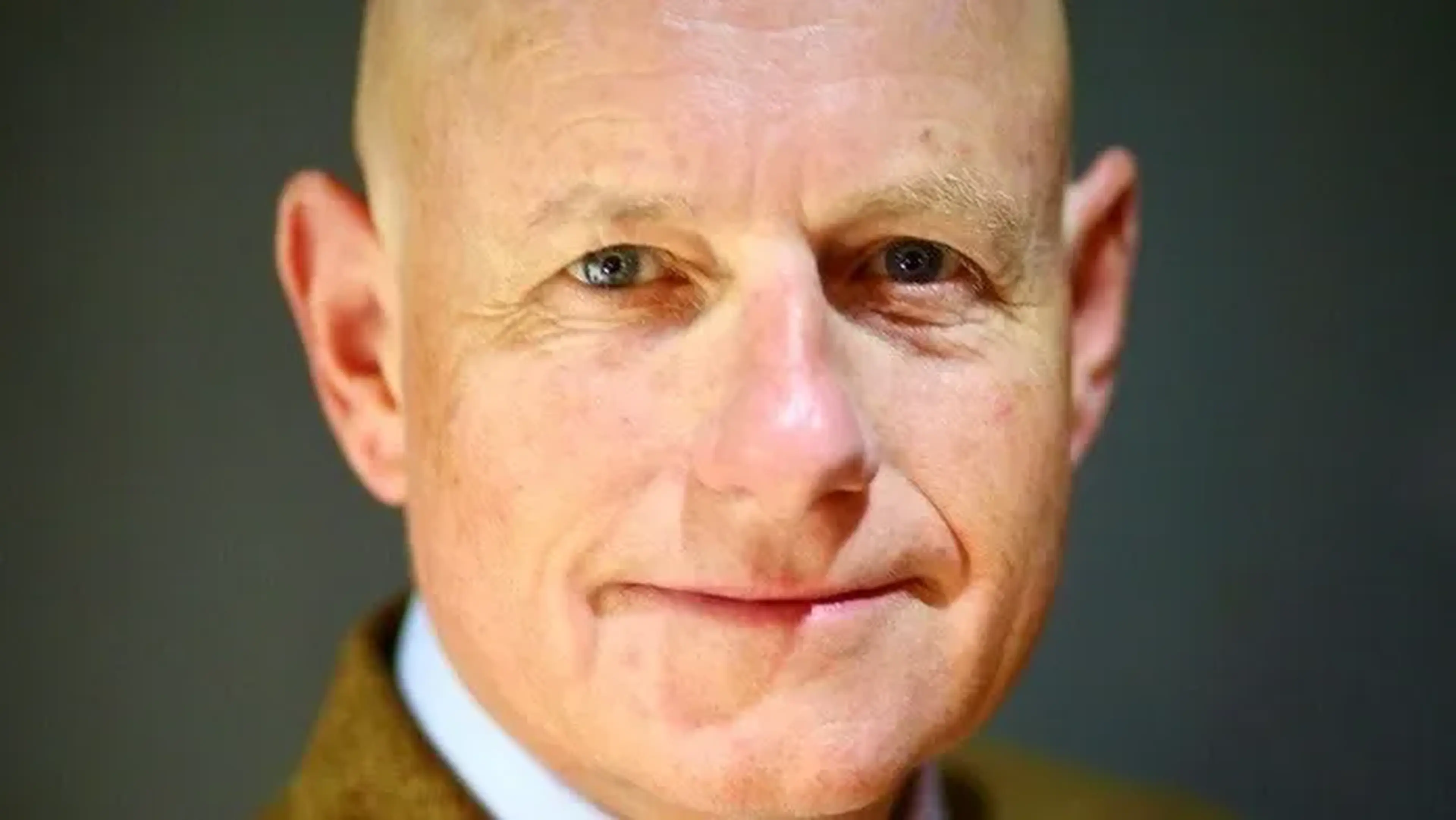 A bald man with fair skin, wearing a brown jacket, looking directly at the camera with a neutral expression against a dark background.