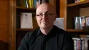 Bald man with glasses in a black shirt, seated in a wooden book-lined room, looking at the camera.