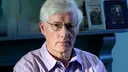 Man with white hair and glasses in a purple shirt sits in a room, with blurry framed photos on a fireplace in the background.