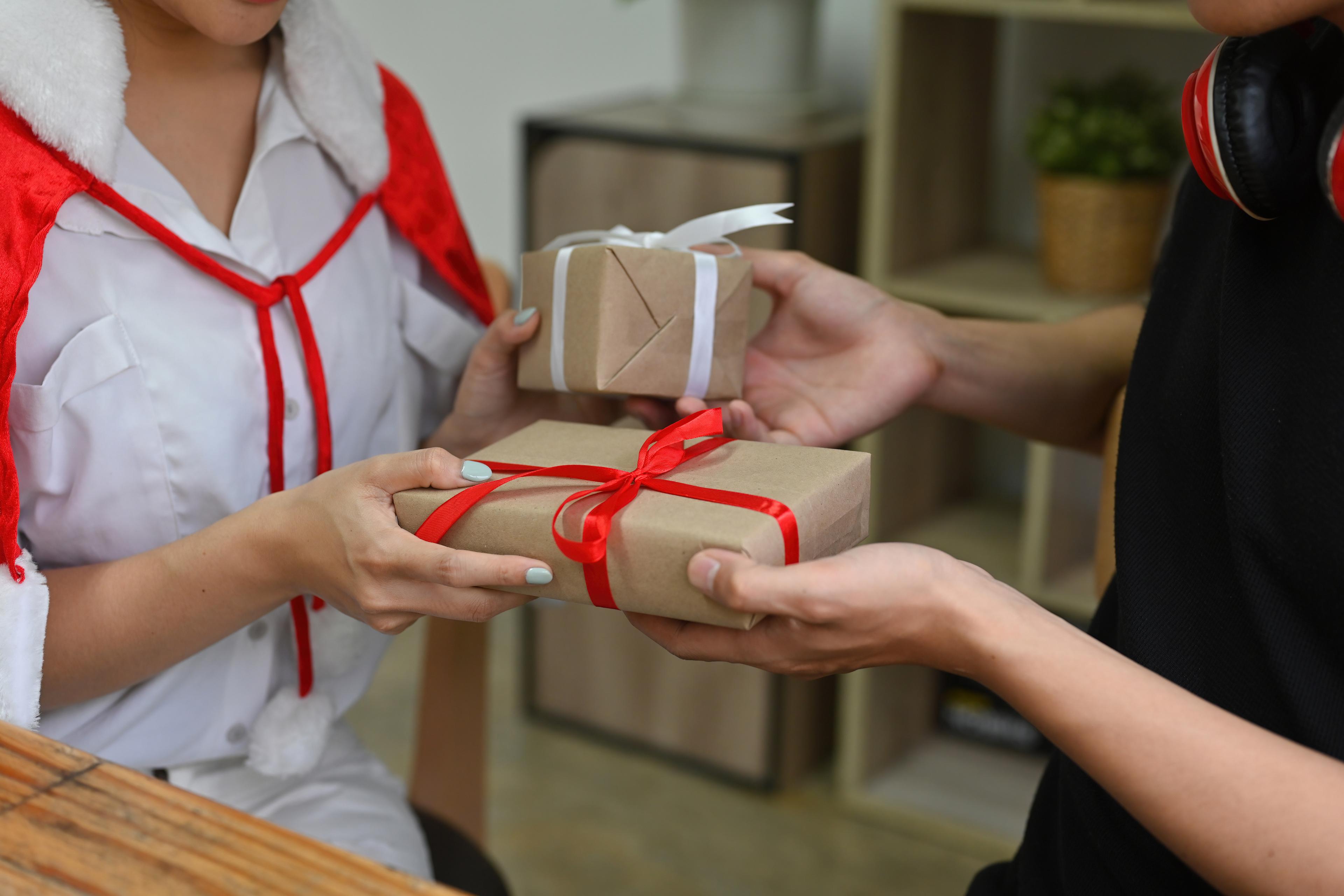 Two people exchanging wrapped gifts; one is dressed in a festive red and white outfit, the other in black with headphones around the neck.