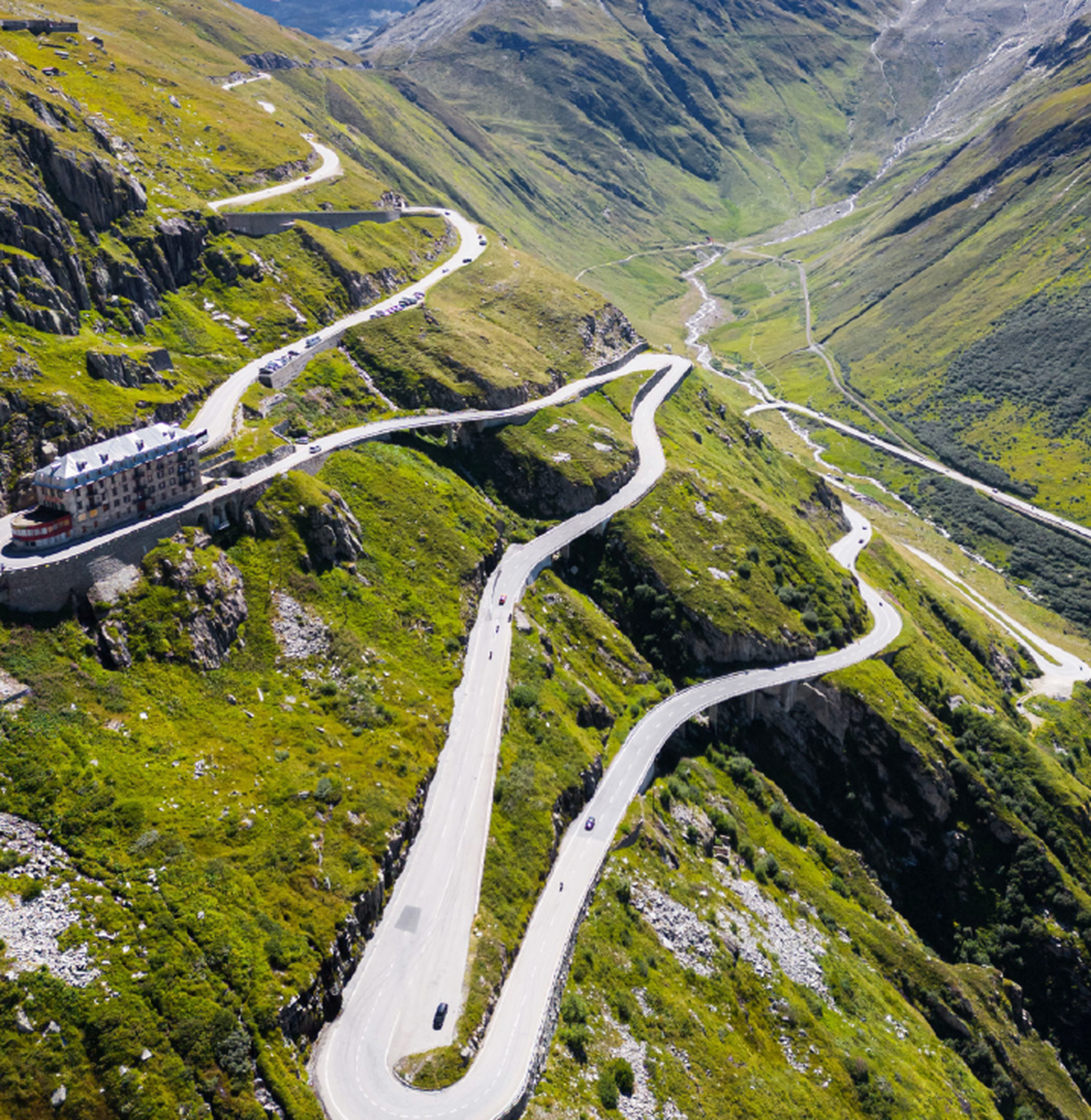 Aerial view of a winding mountain road with sharp curves, surrounded by lush green hillsides and a distant valley.
