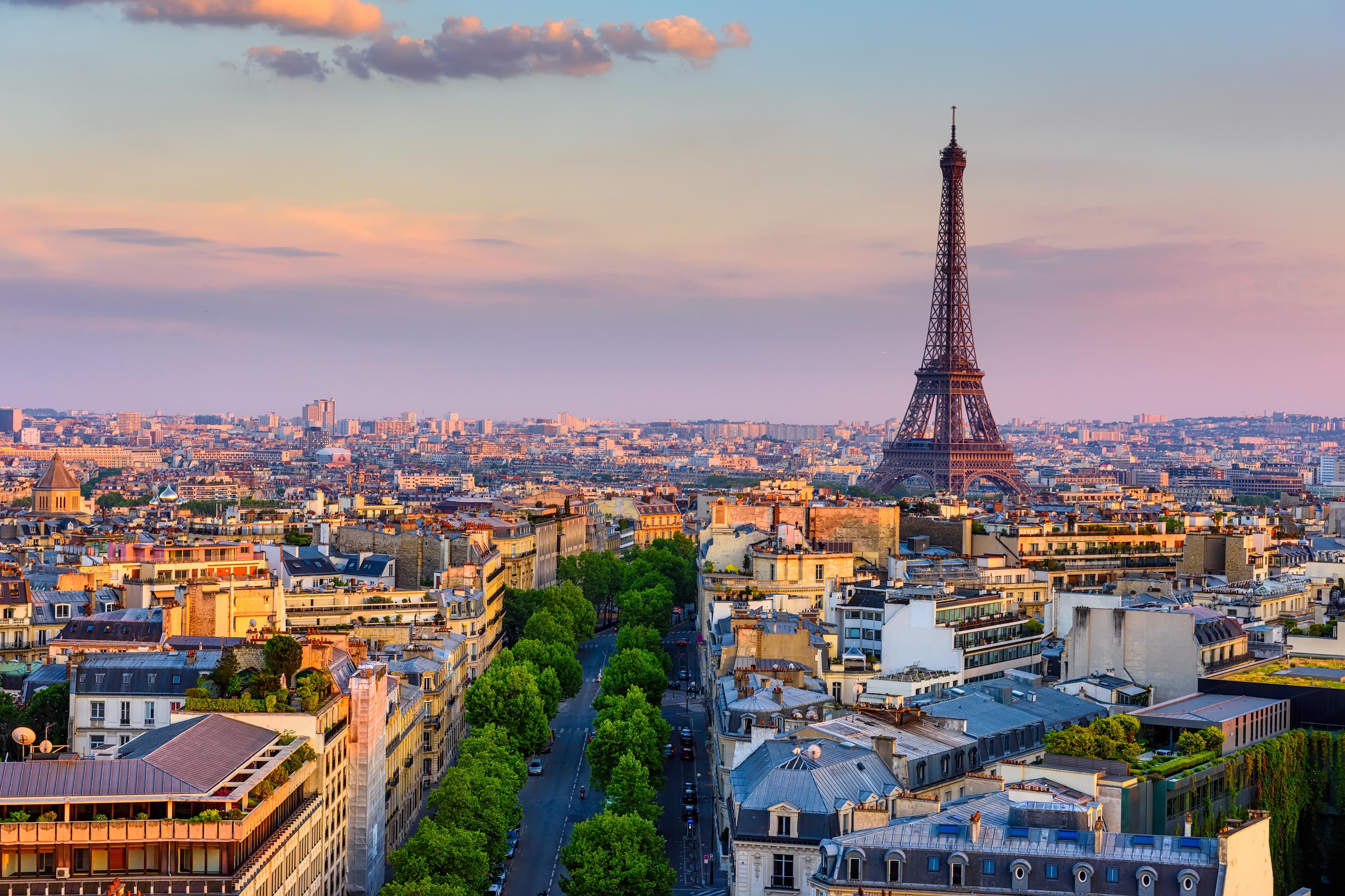 Aerial view of Paris at sunset, featuring the Eiffel Tower and tree-lined streets with historical buildings.