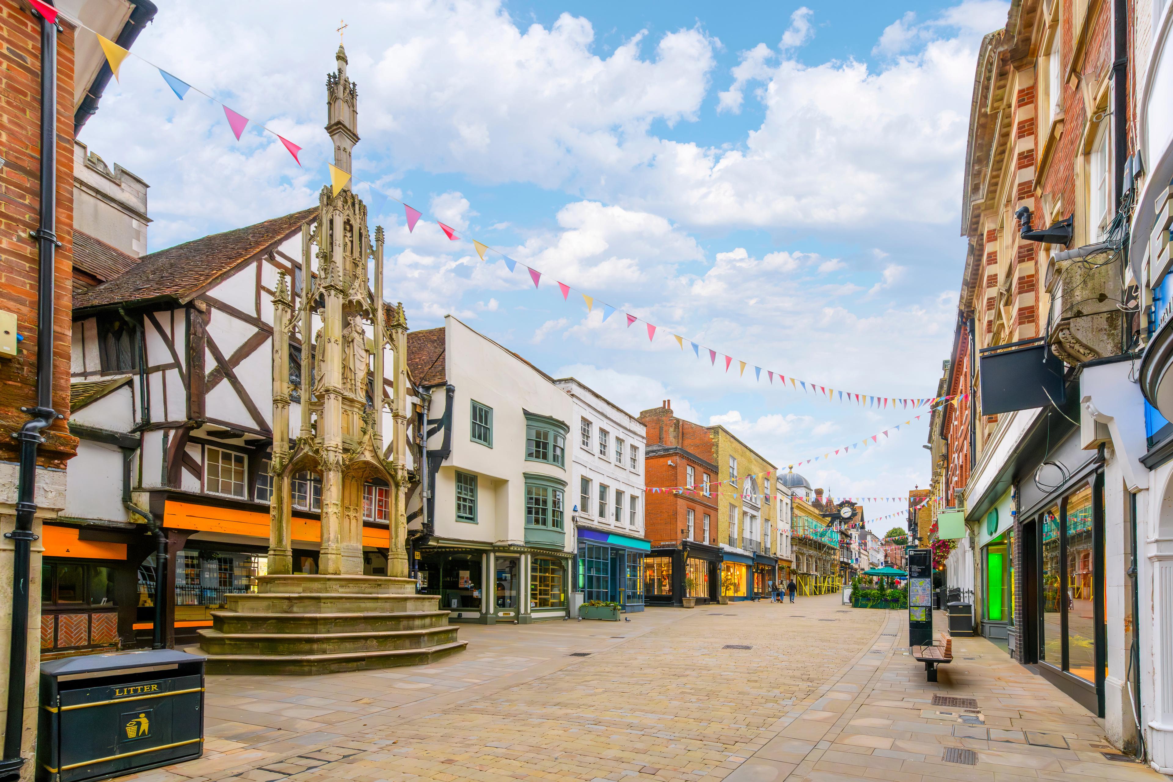 A picturesque street scene with historic buildings, a stone monument, and colorful bunting under a partly cloudy sky.
