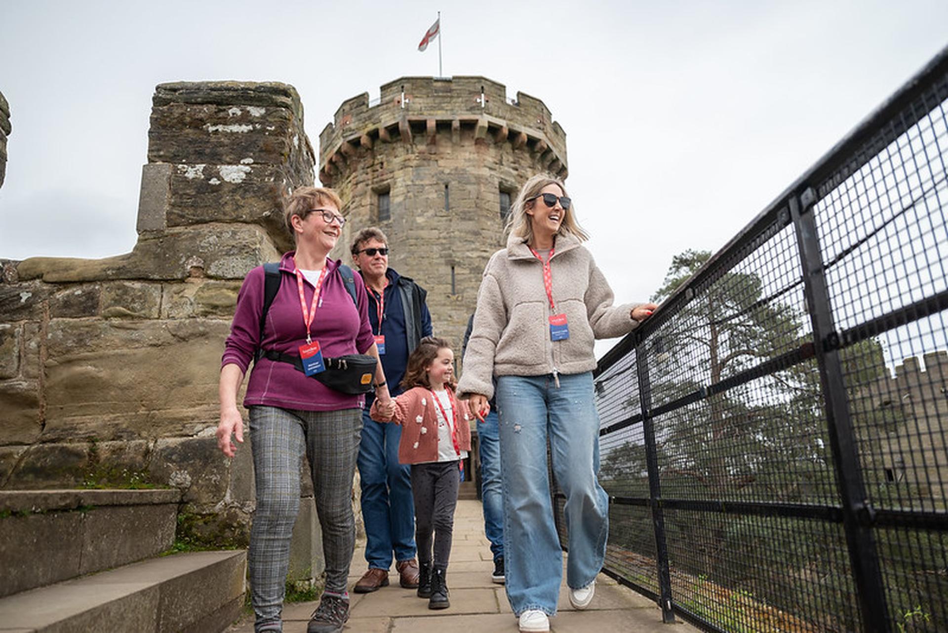 Family walking along a castle wall path on a cloudy day, with a stone tower in the background. They appear happy and engaged in conversation.