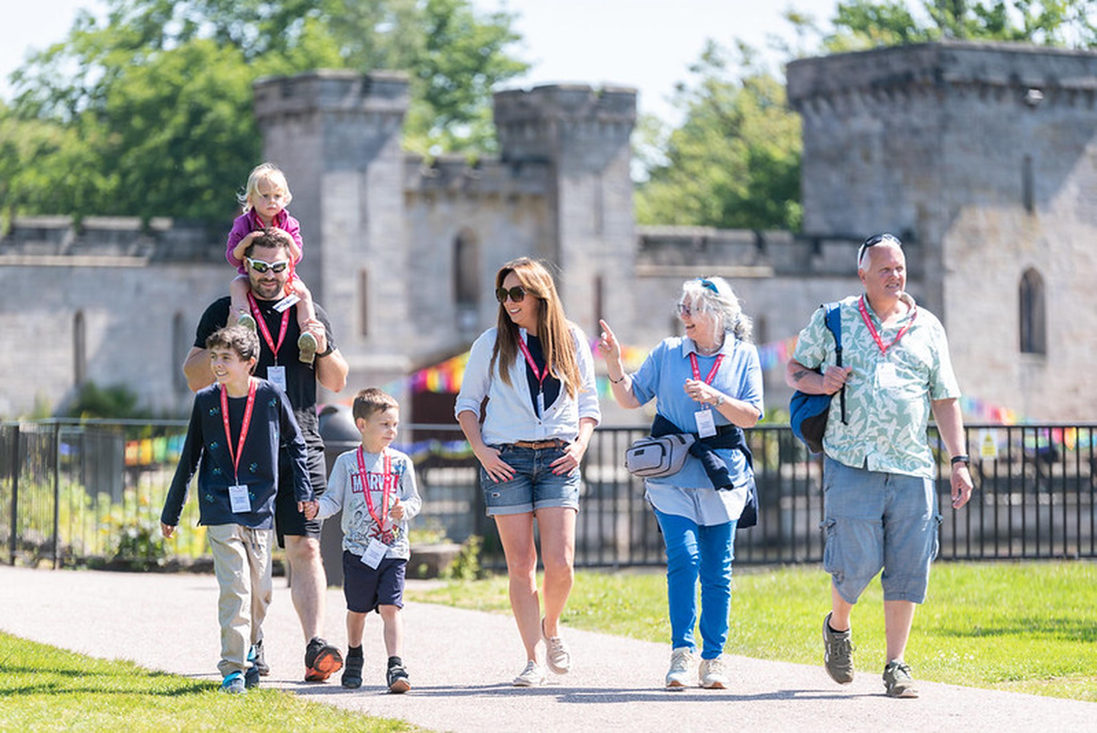 A group of people, including children, walking outside near a castle, wearing lanyards on a sunny day.