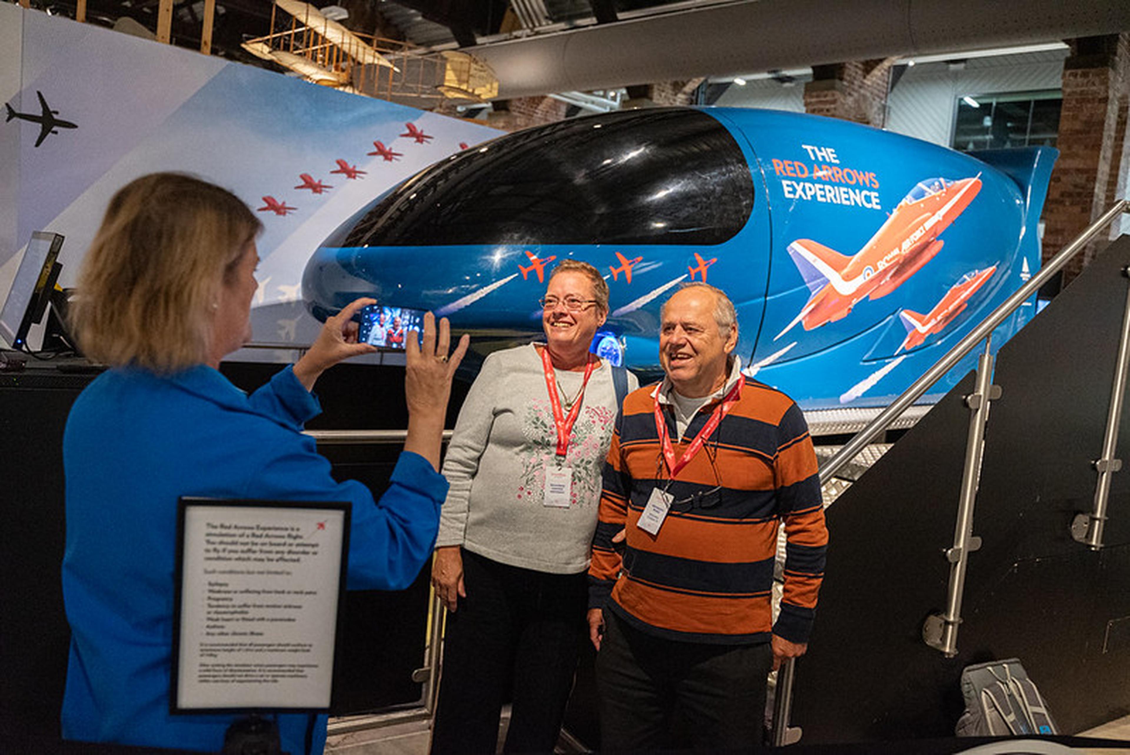 A woman photographs a smiling couple in front of "The Red Arrows Experience" simulator