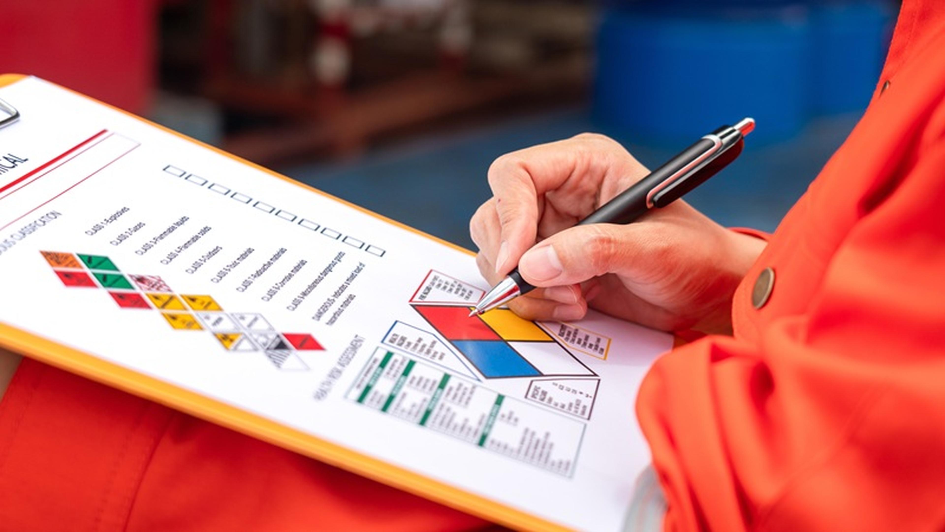 Person in orange uniform fills out a safety risk assessment form on a clipboard, featuring a hazard diamond chart and classifications.