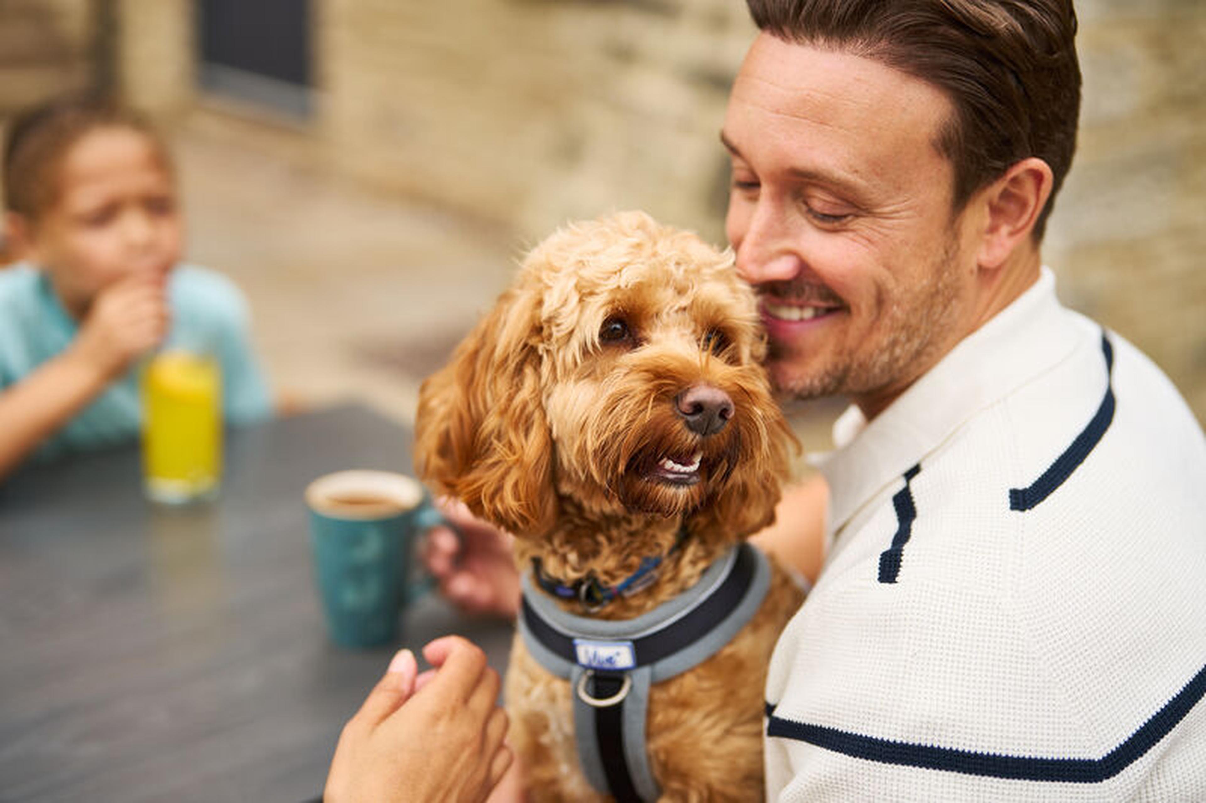 Man smiling at a small, fluffy dog on his lap at an outdoor table, with a child in the background drinking from a glass.