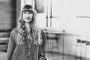 Black and white photo of a person with long wavy hair and bangs, wearing a button-up shirt, standing in a well-lit room.