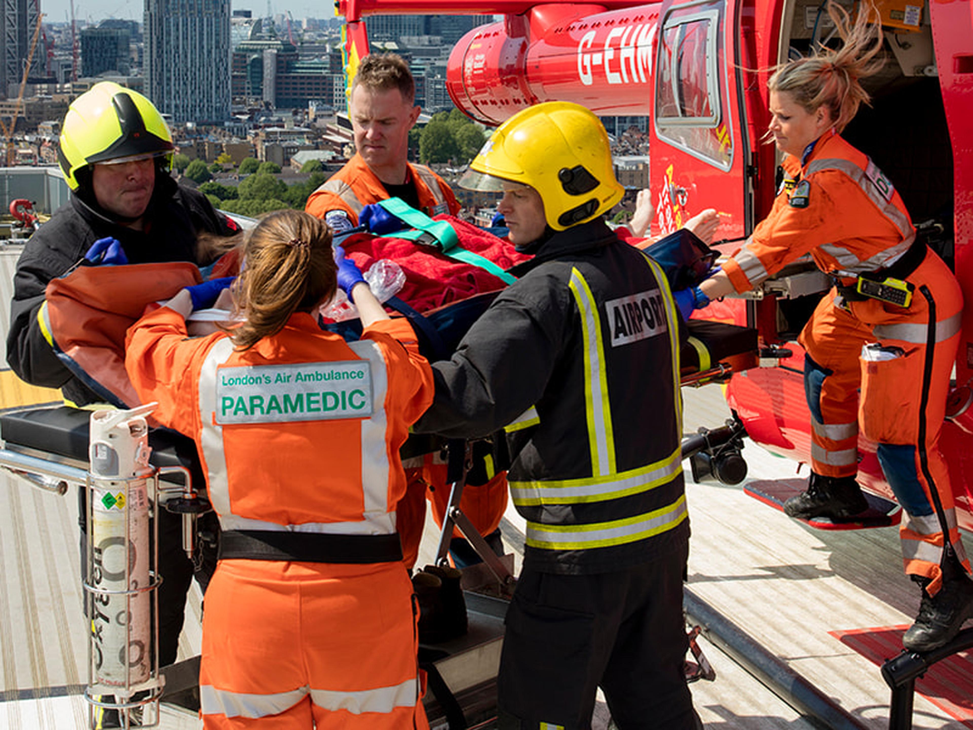 Paramedics loading a patient onto a stretcher from a red helicopter on a rooftop in a cityscape setting.