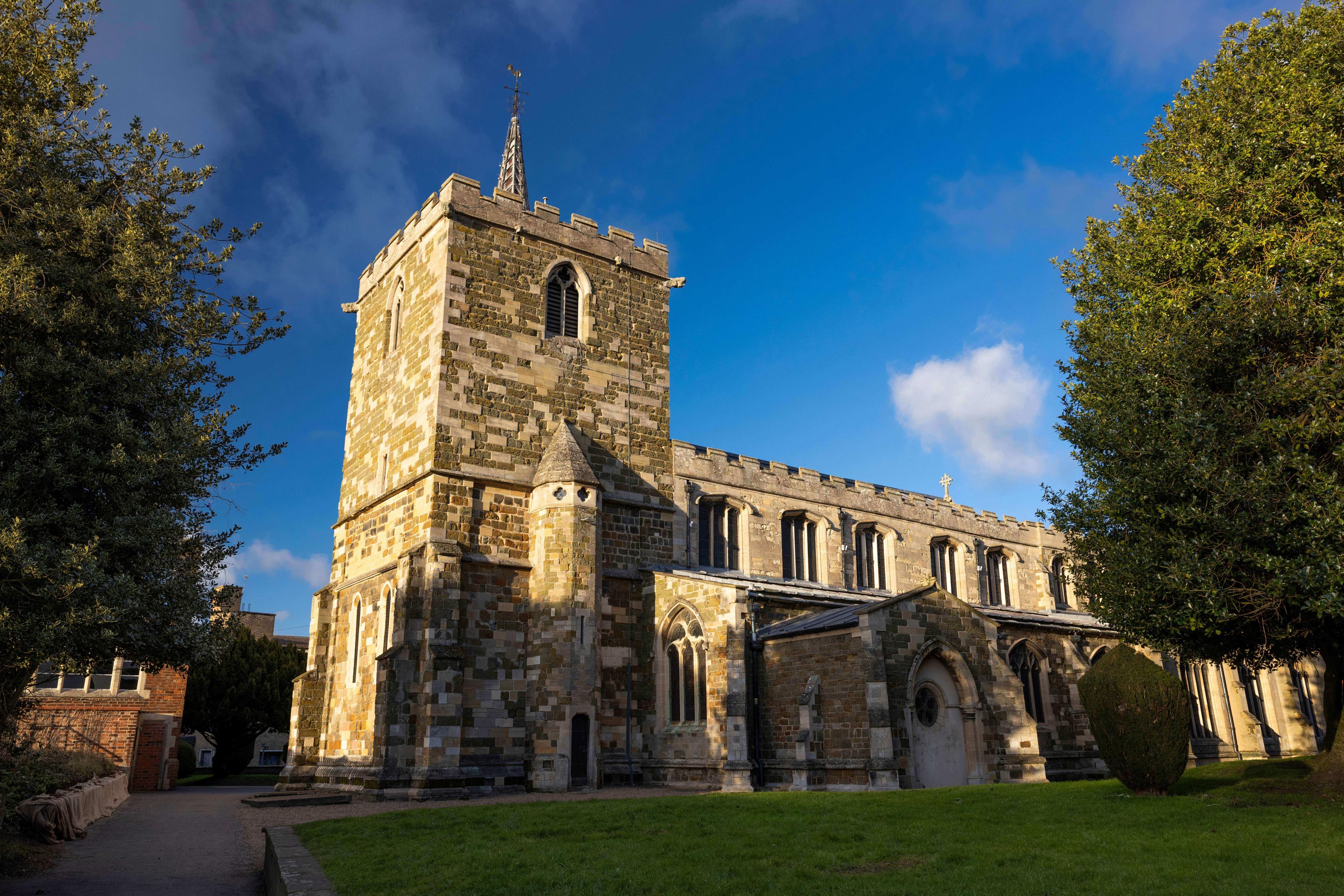 Historic stone church with a square tower under a blue sky, surrounded by green trees and grass.