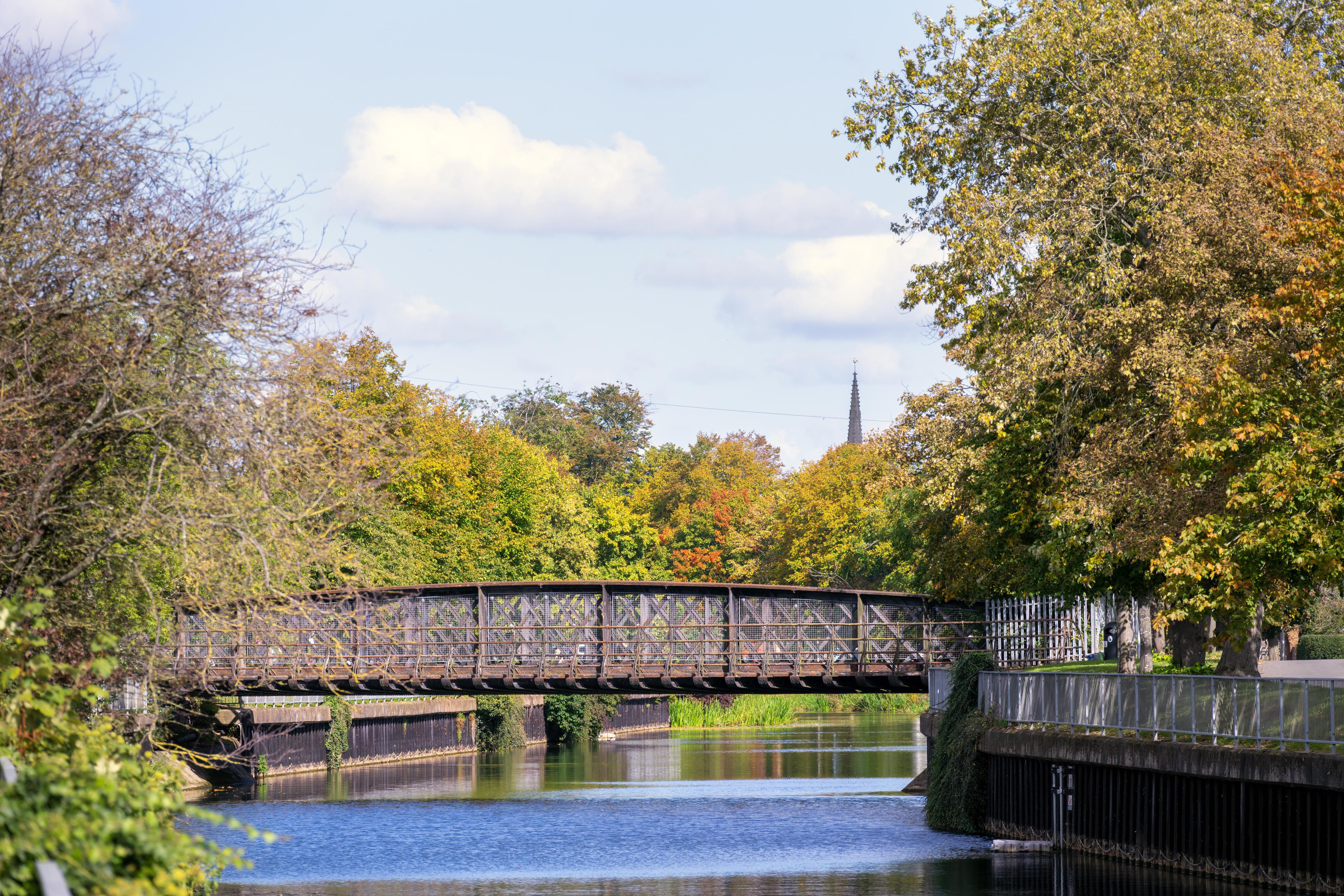 A metal bridge spans a calm river, surrounded by lush green and autumnal trees under a partly cloudy sky.