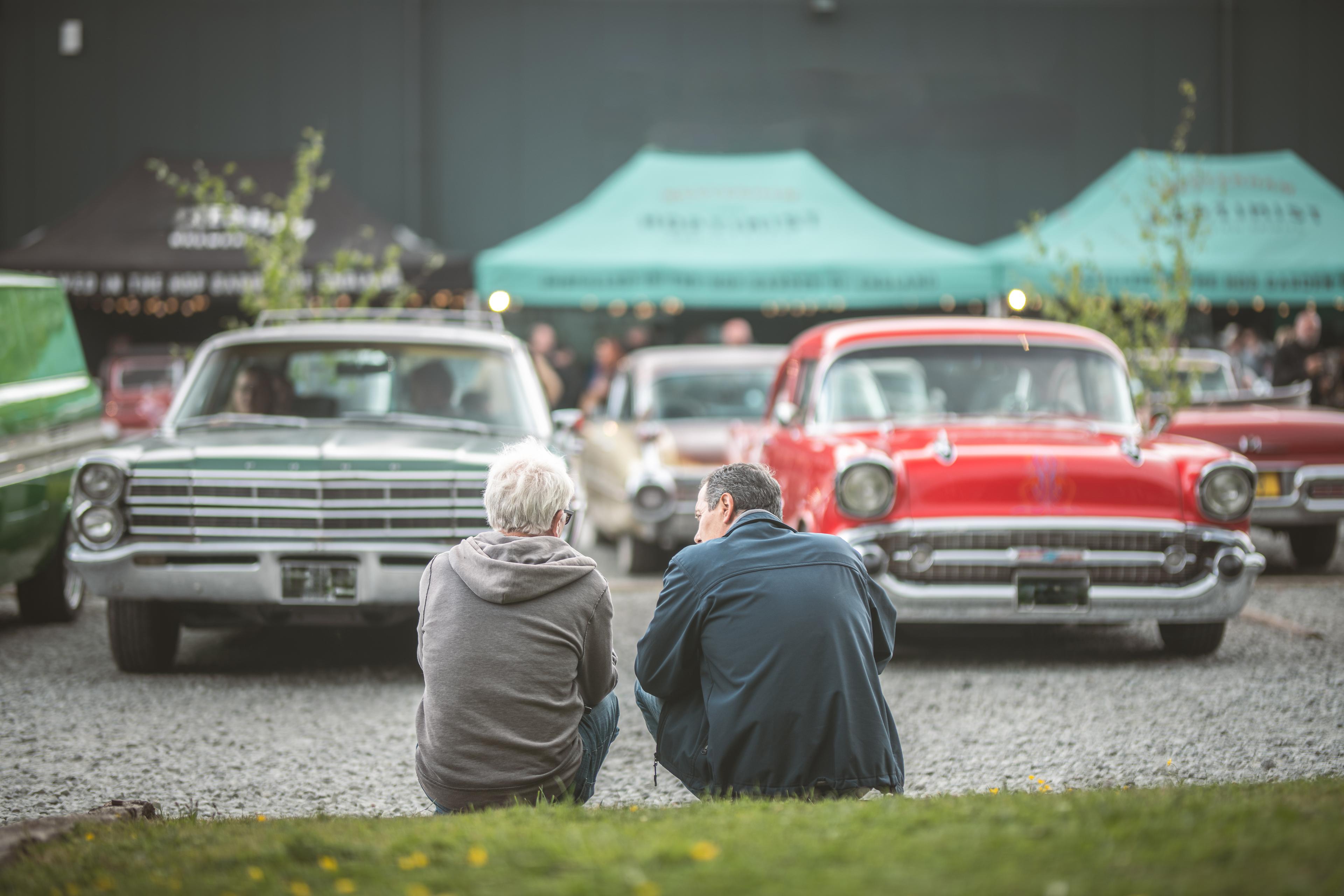 Two men sit on a curb, facing a lineup of vintage cars, including a green and a red car, with event tents in the background.