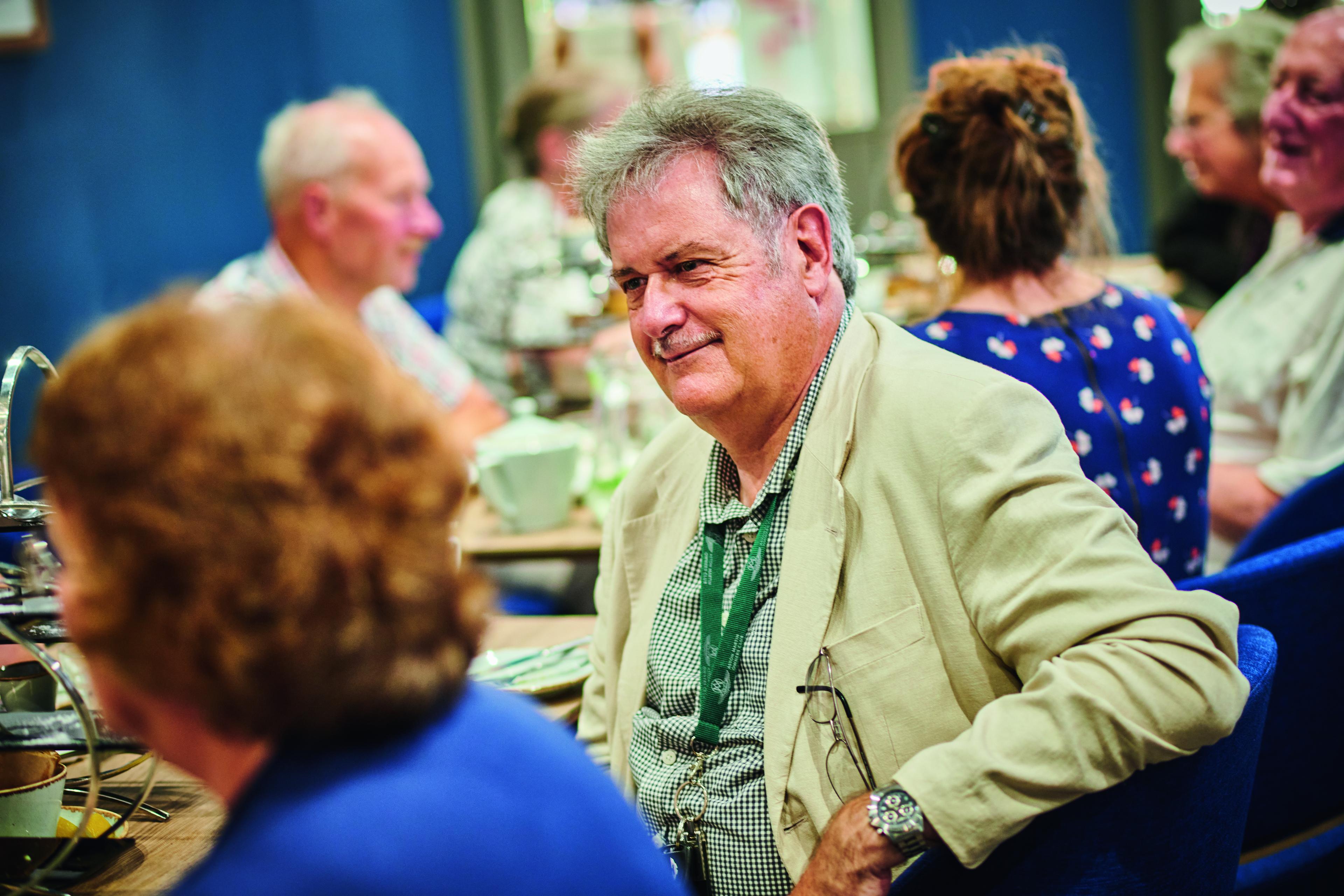 A man in a beige jacket is sitting at a dining table, engaged in conversation with others, in a cozy, social setting.