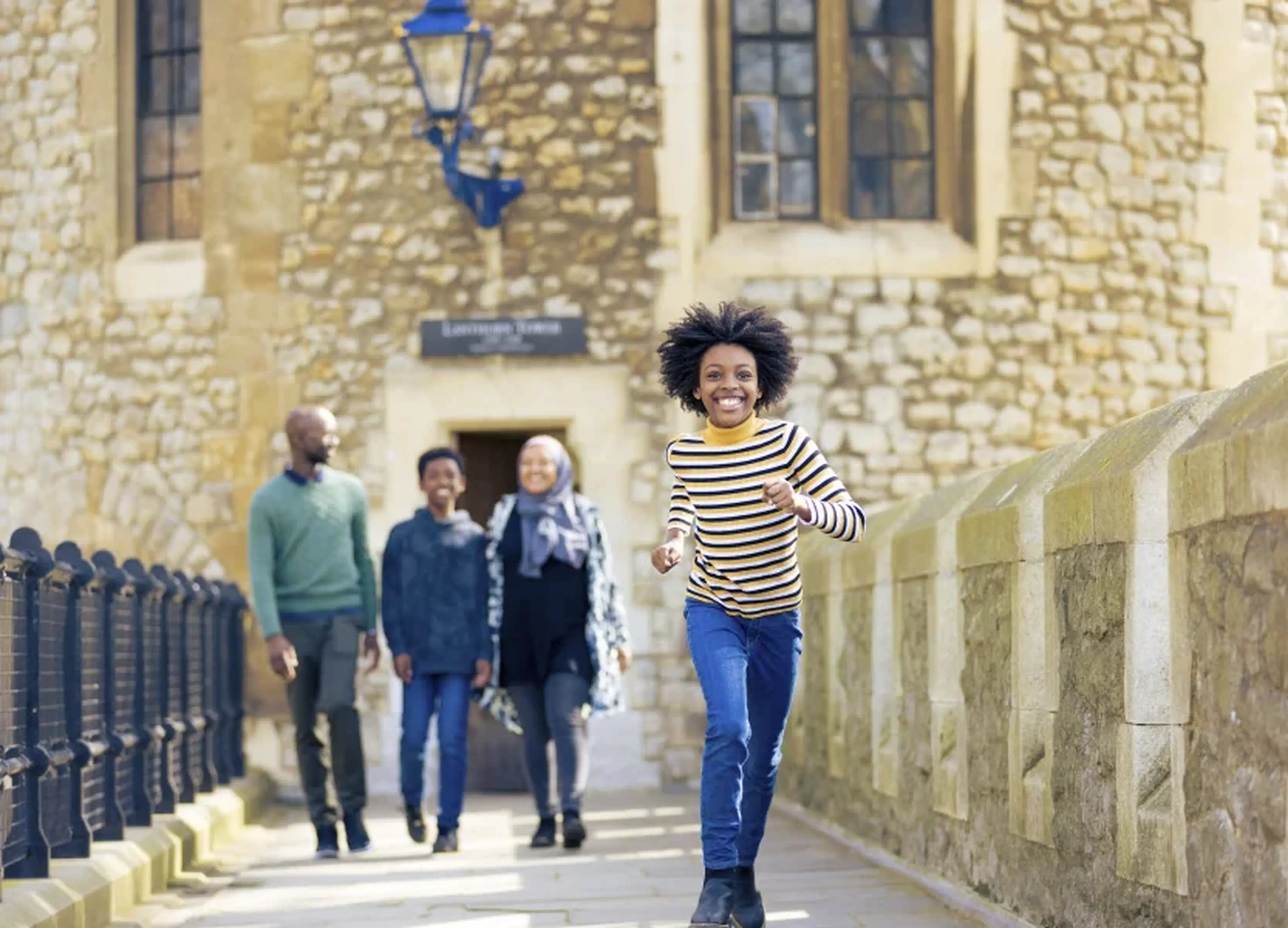 A joyful child runs towards the camera while three adults walk behind, set against a historic stone building with windows and a lantern.