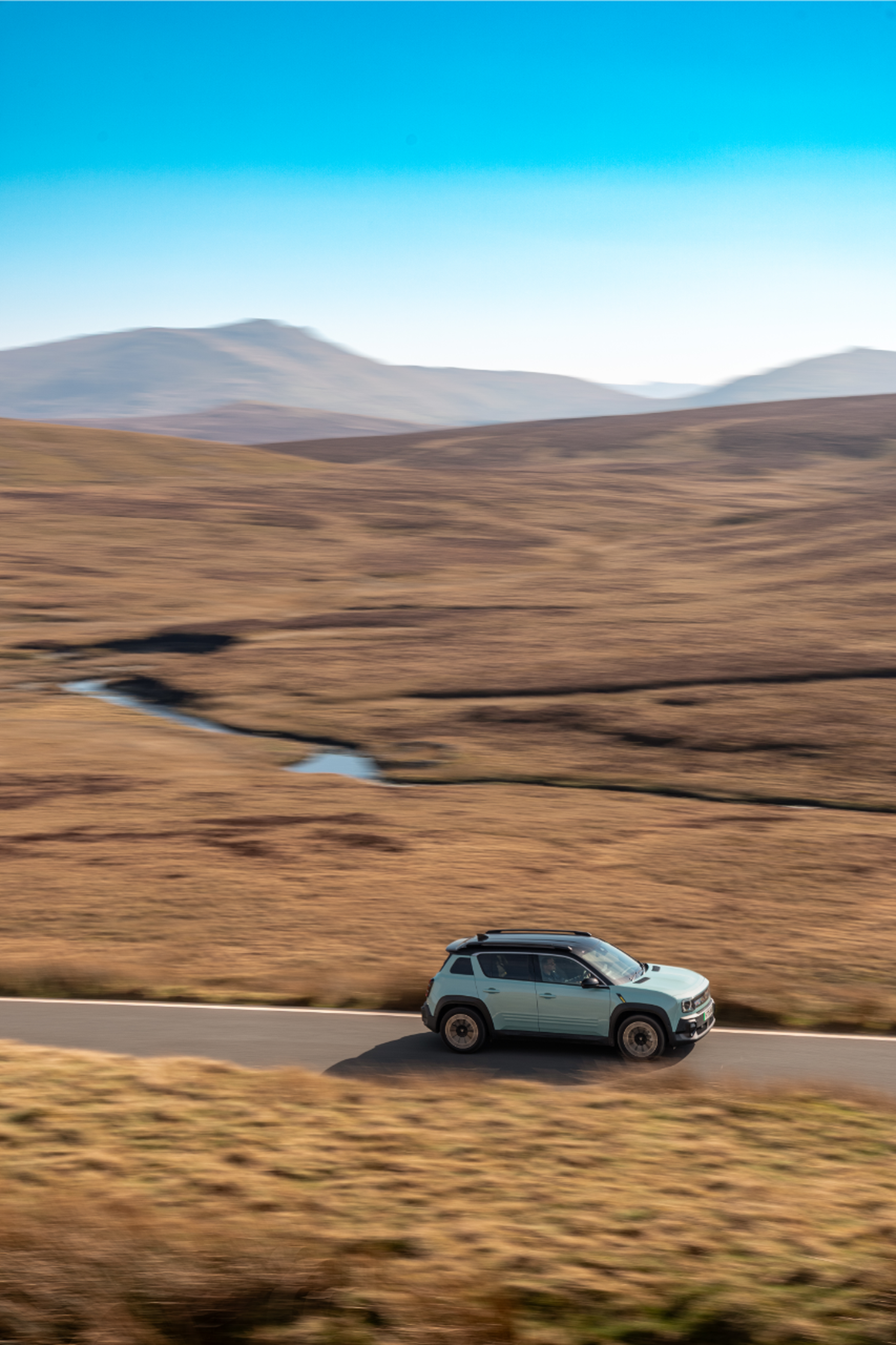 A light blue SUV drives on a winding road through a vast, brown barren landscape with distant mountains under a clear blue sky.