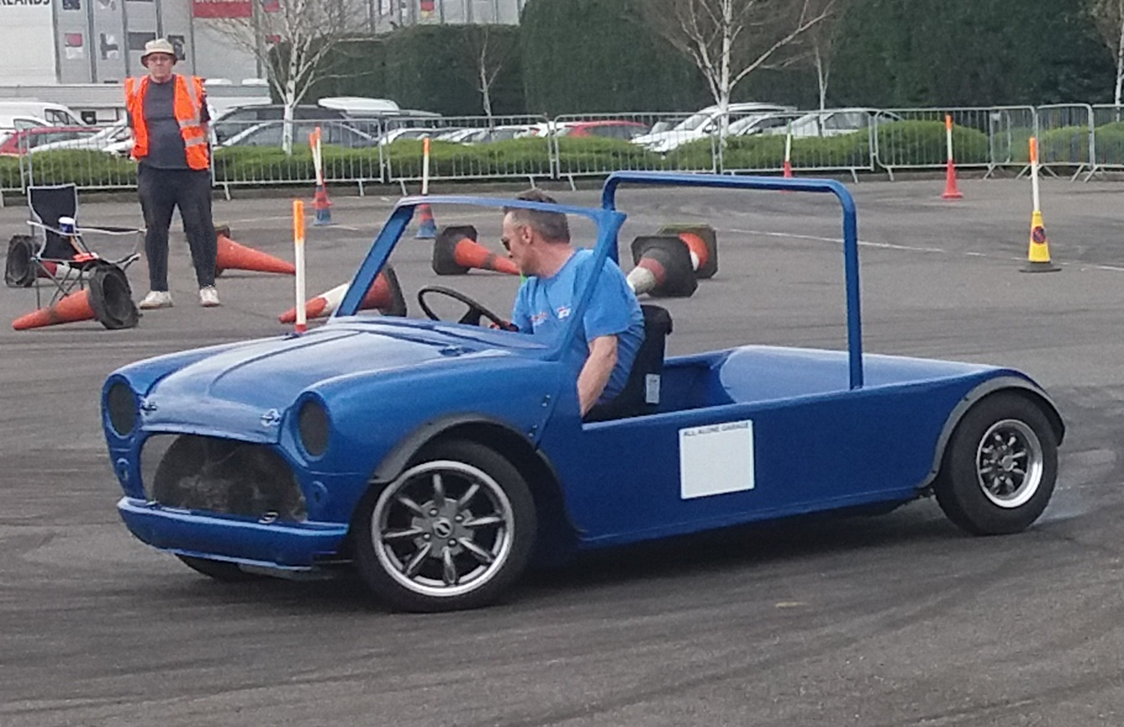 A man driving a small, blue, roofless car in a parking area