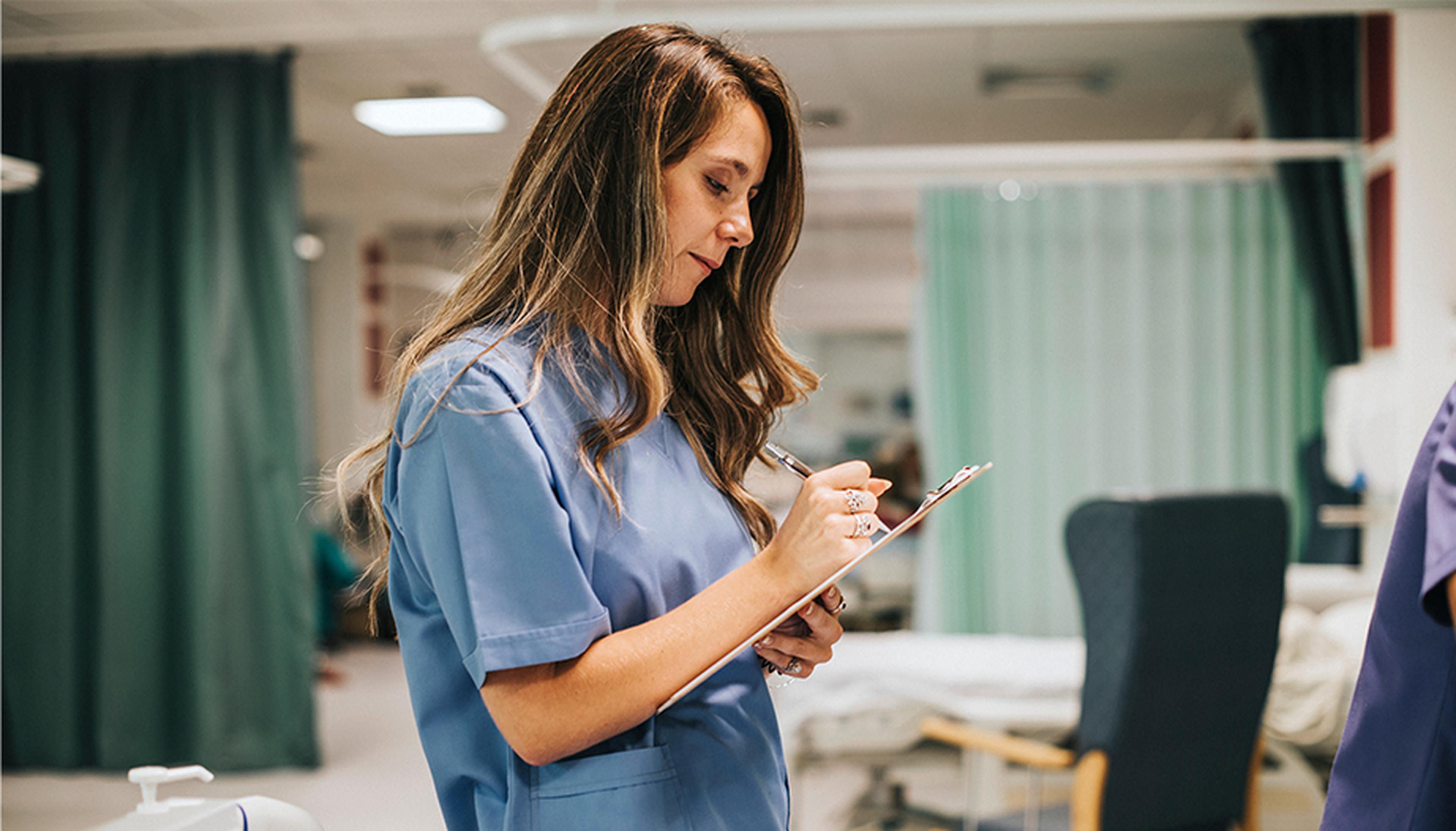 Healthcare professional in blue scrubs writing on a clipboard in a hospital ward, with medical equipment and curtains in the background.