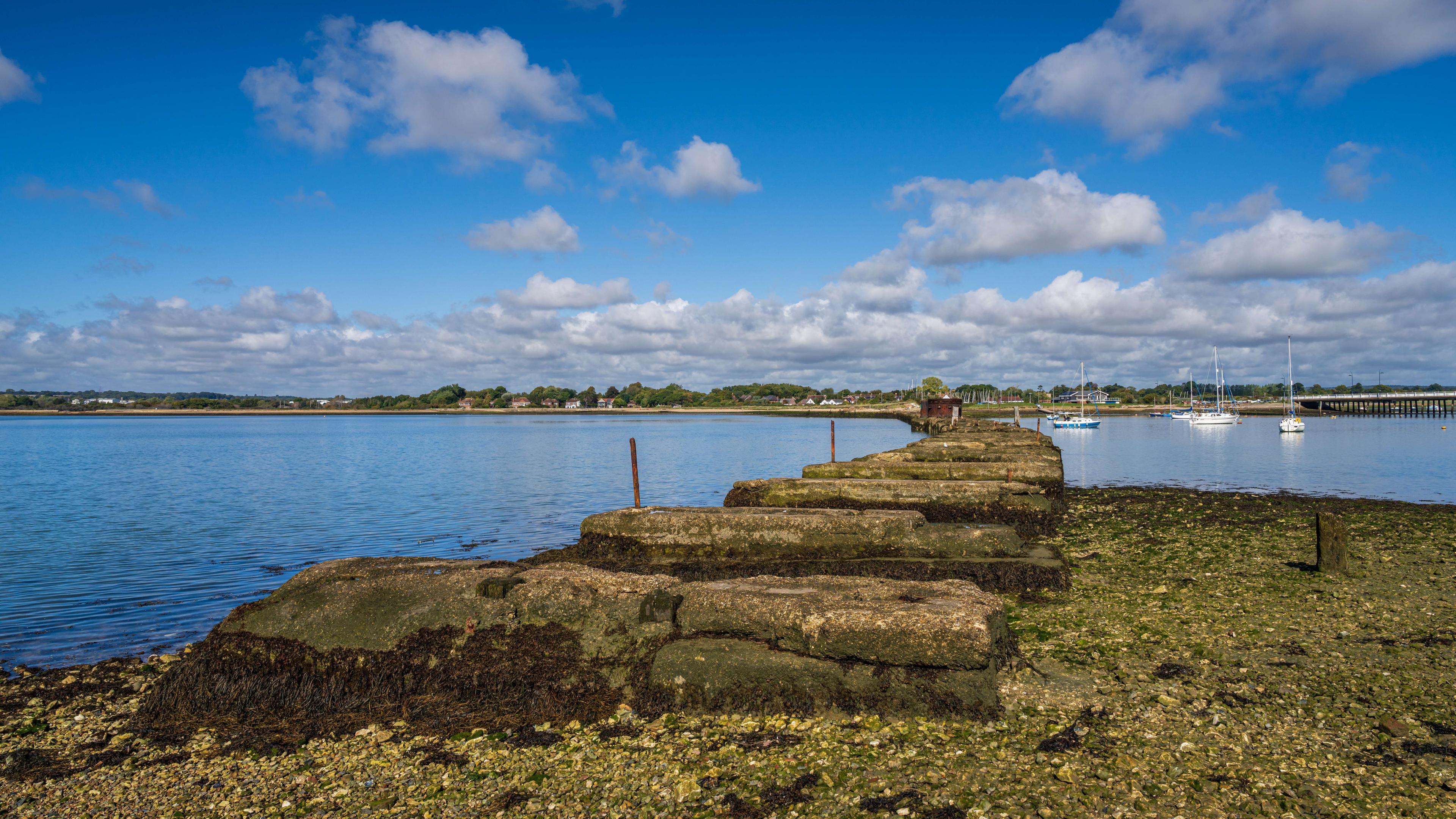 Seaweed-covered stone groynes stretch across a calm tidal shore toward a harbor of sailboats, beneath a bright blue sky; the scene feels peaceful.