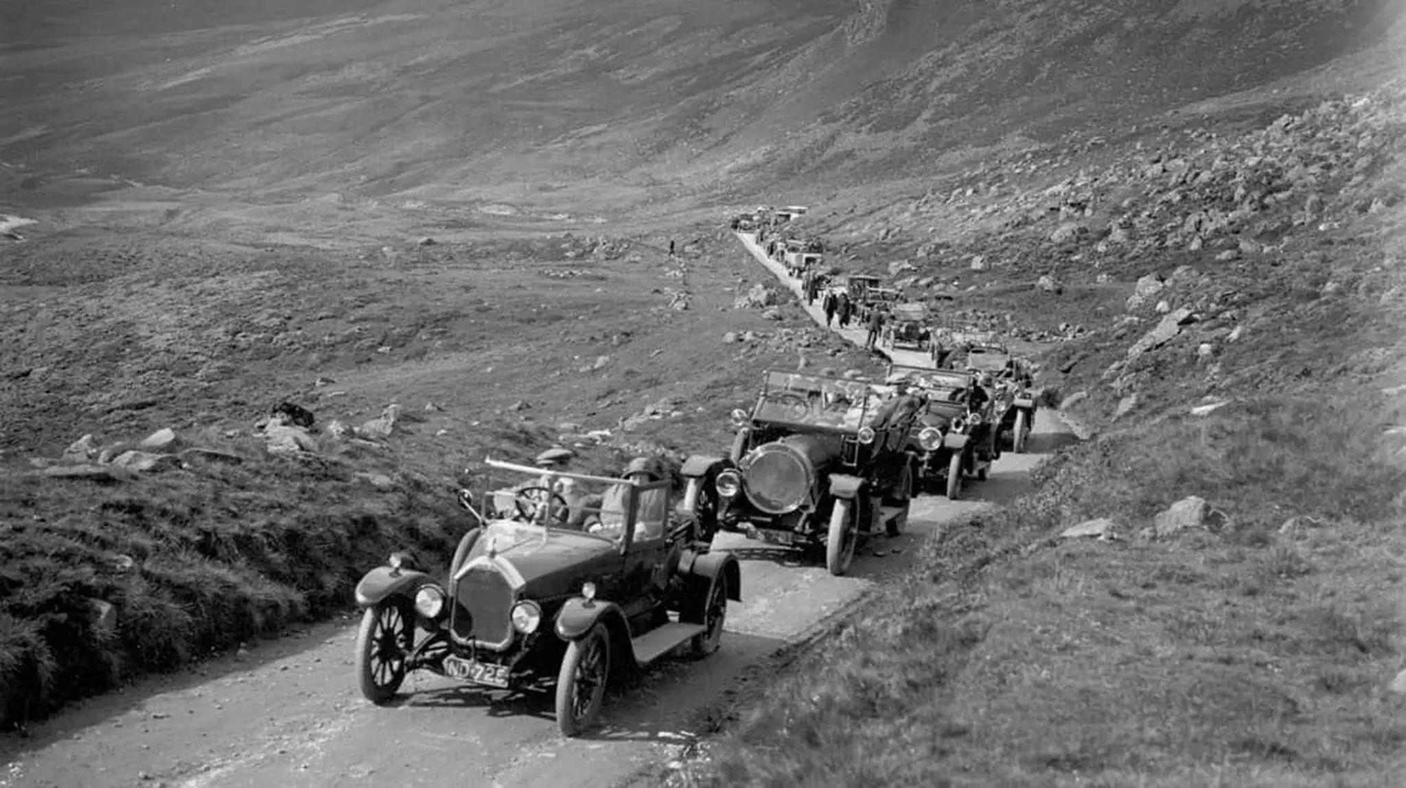 A line of vintage cars travels up a narrow, winding road on a rugged hillside