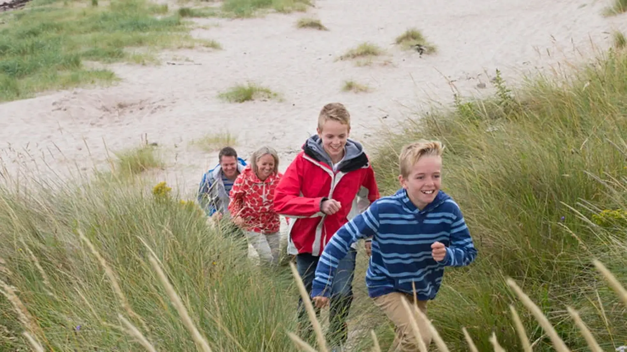 A family of four, wearing jackets, joyfully climbs a sandy dune